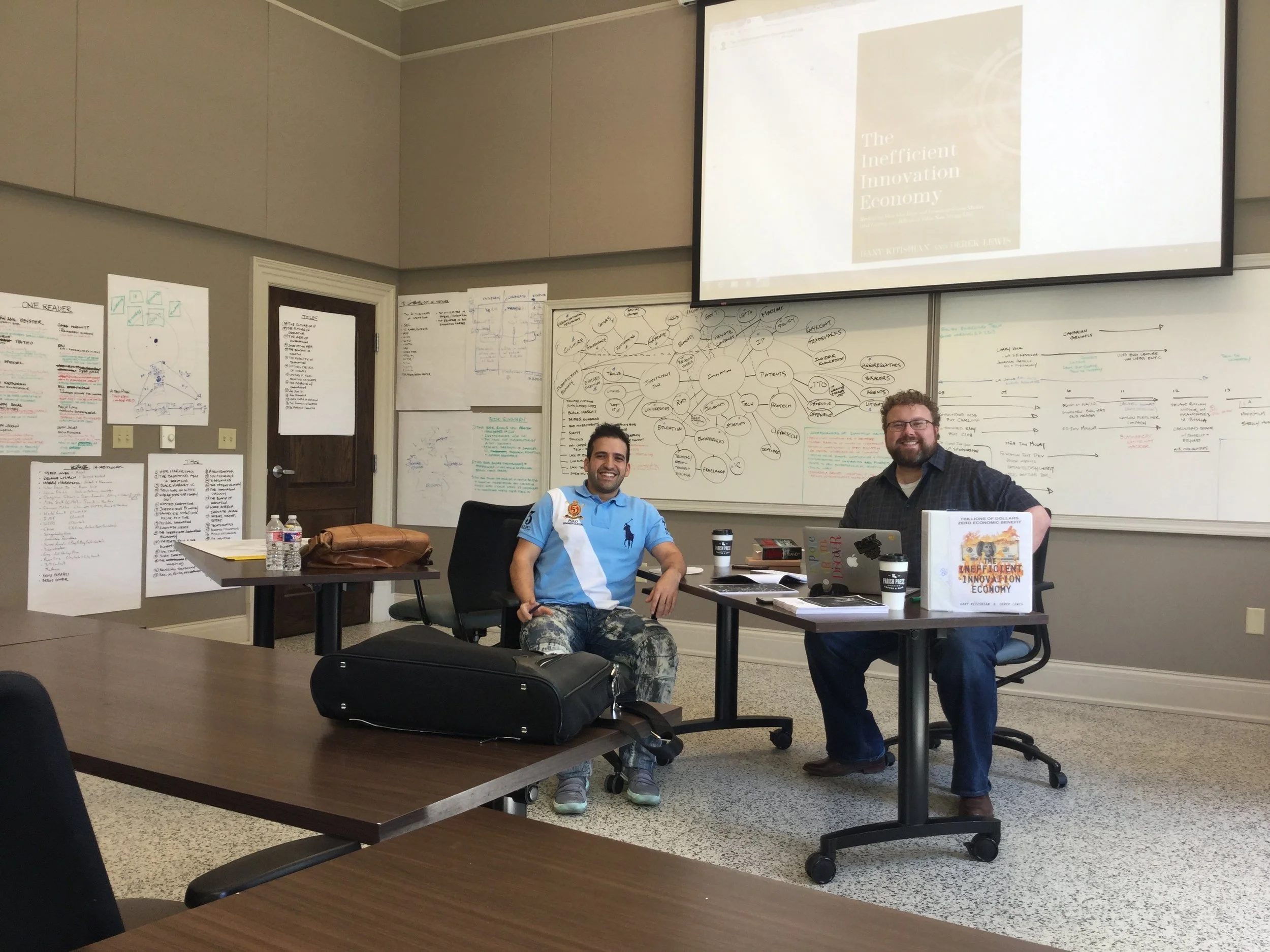 Two men smiling and sitting in a classroom or meeting room, with whiteboards and diagrams on the wall behind them. One man is wearing a light blue polo shirt and the other is wearing a black shirt, with a laptop, books, and coffee cups on the table in front of them.