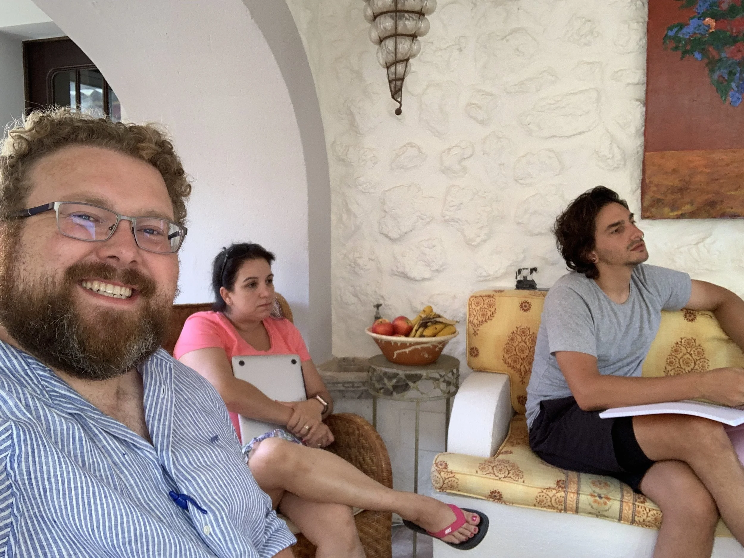 Three people sitting in a living room. The man on the left is smiling at the camera, wearing glasses and a striped shirt. The woman in the middle is holding a tablet and looking to her left, wearing a pink top. The man on the right is sitting on the arm of a sofa, looking to his left, wearing a gray t-shirt and black shorts, with a notebook on his lap. There is a bowl of fruit on a table behind them against a white textured wall.