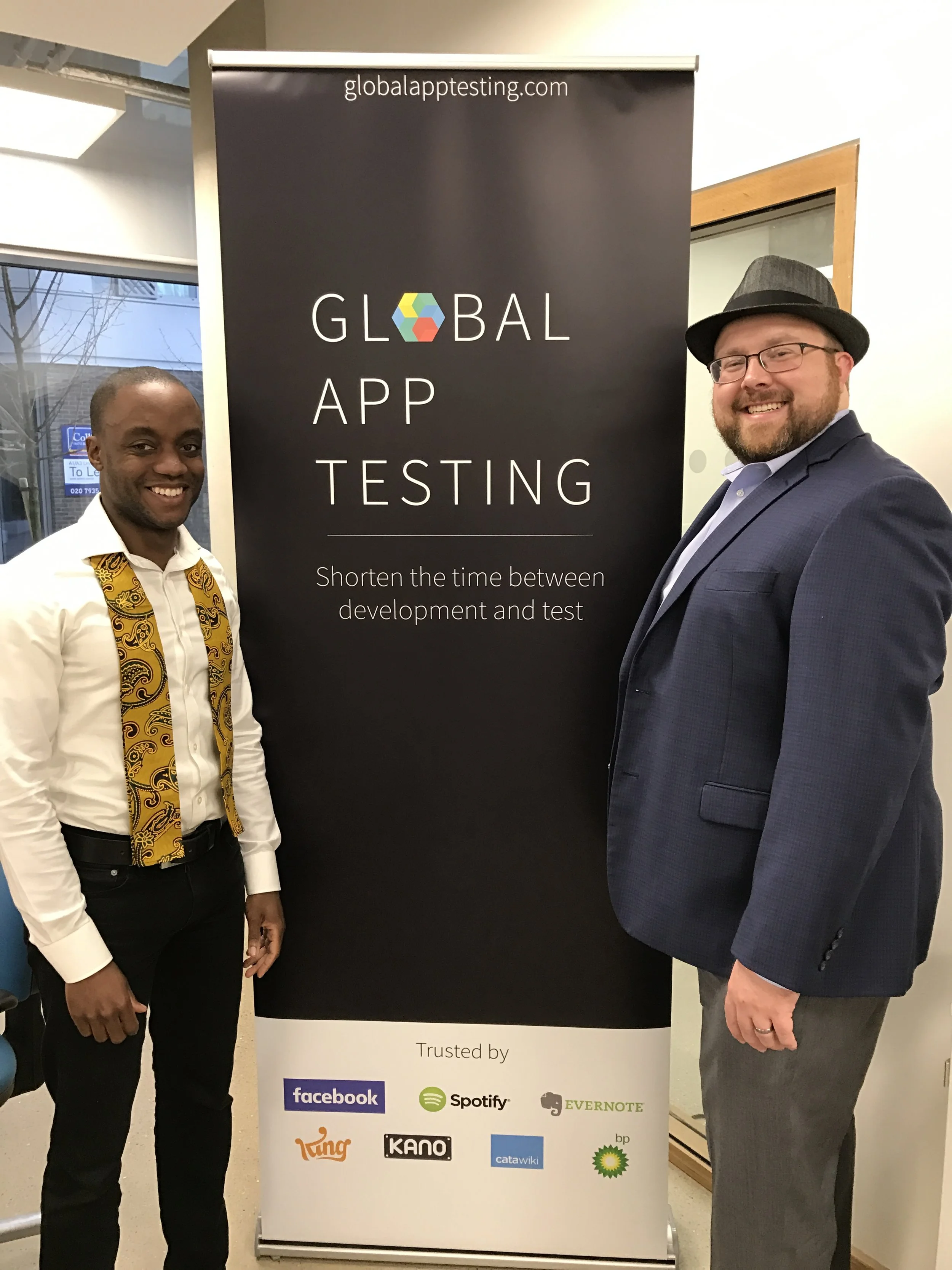 Two men standing beside a black vertical banner that reads "GLOBAL APP TESTING" and "Shorten the time between development and test" with logos of Facebook, Spotify, Evernote, Turing, Kano, Catawiki, and BP at the bottom. The man on the left is wearing a white shirt with a yellow patterned tie, and the man on the right is wearing a blue blazer, glasses, and a hat.