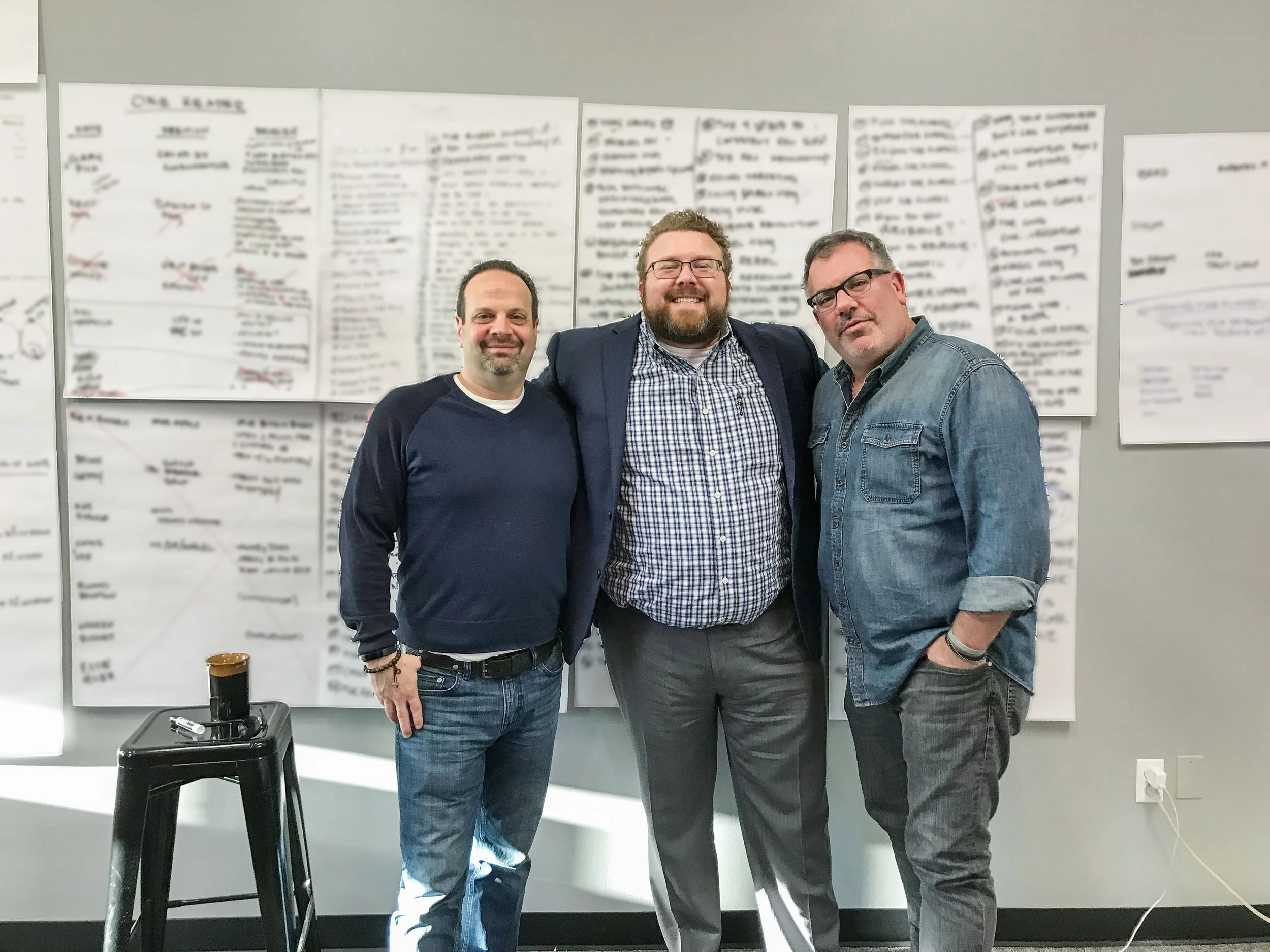 Three men standing together indoors, smiling, with a blurred whiteboard or large paper sheets with notes behind them.