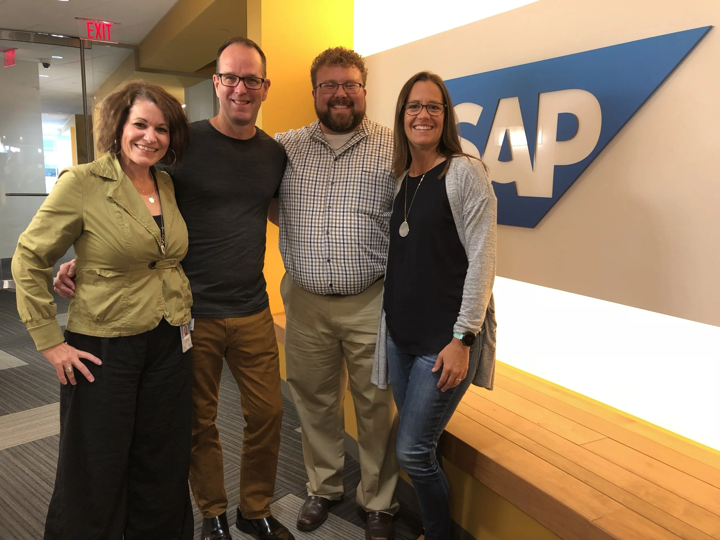 Four people standing in front of a SAP sign, smiling for the photo in an office lobby.