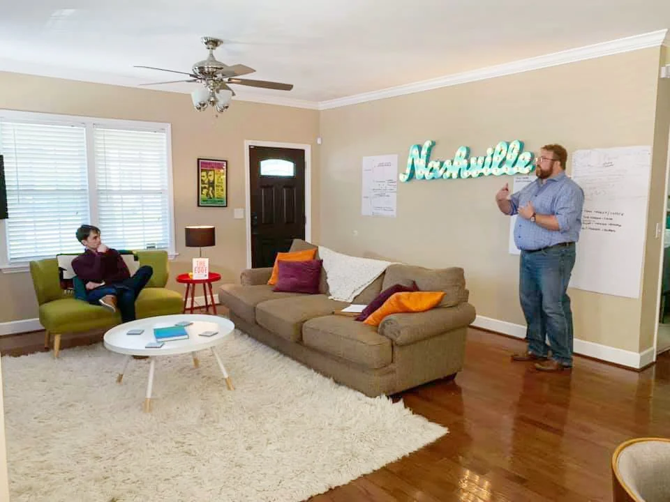 Living room with two people, one sitting on a green chair and one standing near the wall, which has a illuminated 'Nashville' sign. The room has beige walls, a hanging ceiling fan, a window with blinds, a small red side table, a beige sofa with colorful pillows, a white round coffee table, and a white shag rug.
