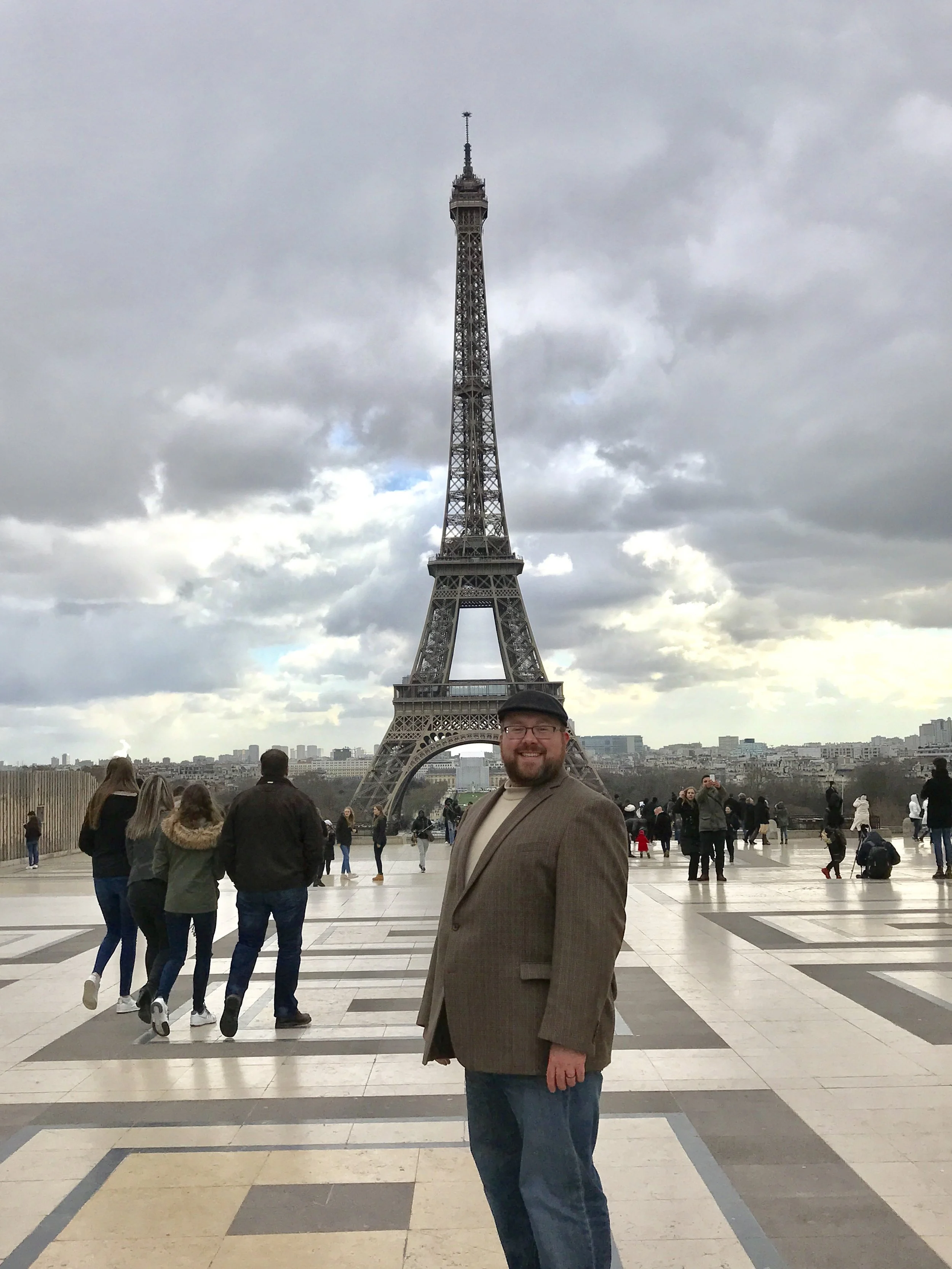 A man smiling in front of the Eiffel Tower in Paris, France, with tourists around him and a cloudy sky overhead.