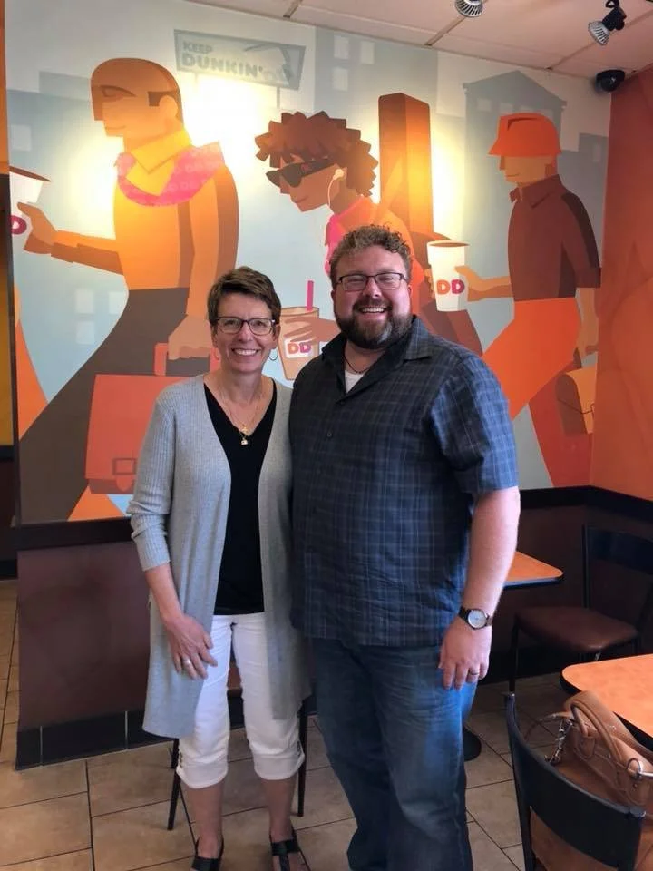 Two people, a woman and a man, standing inside a Dunkin' Donuts restaurant with a colorful wall mural of animated figures enjoying coffee and drinks in the background.