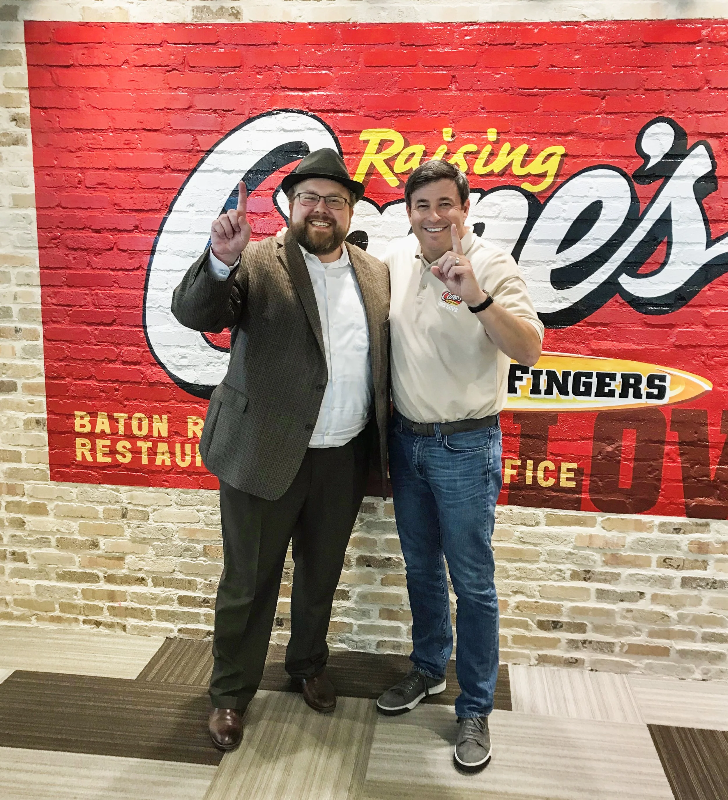 Two men standing in front of a red brick wall with a large sign that reads "Raising Cane's." The man on the left is wearing glasses, a hat, a brown checked blazer, white shirt, and dark trousers. The man on the right is wearing a white polo shirt wit
