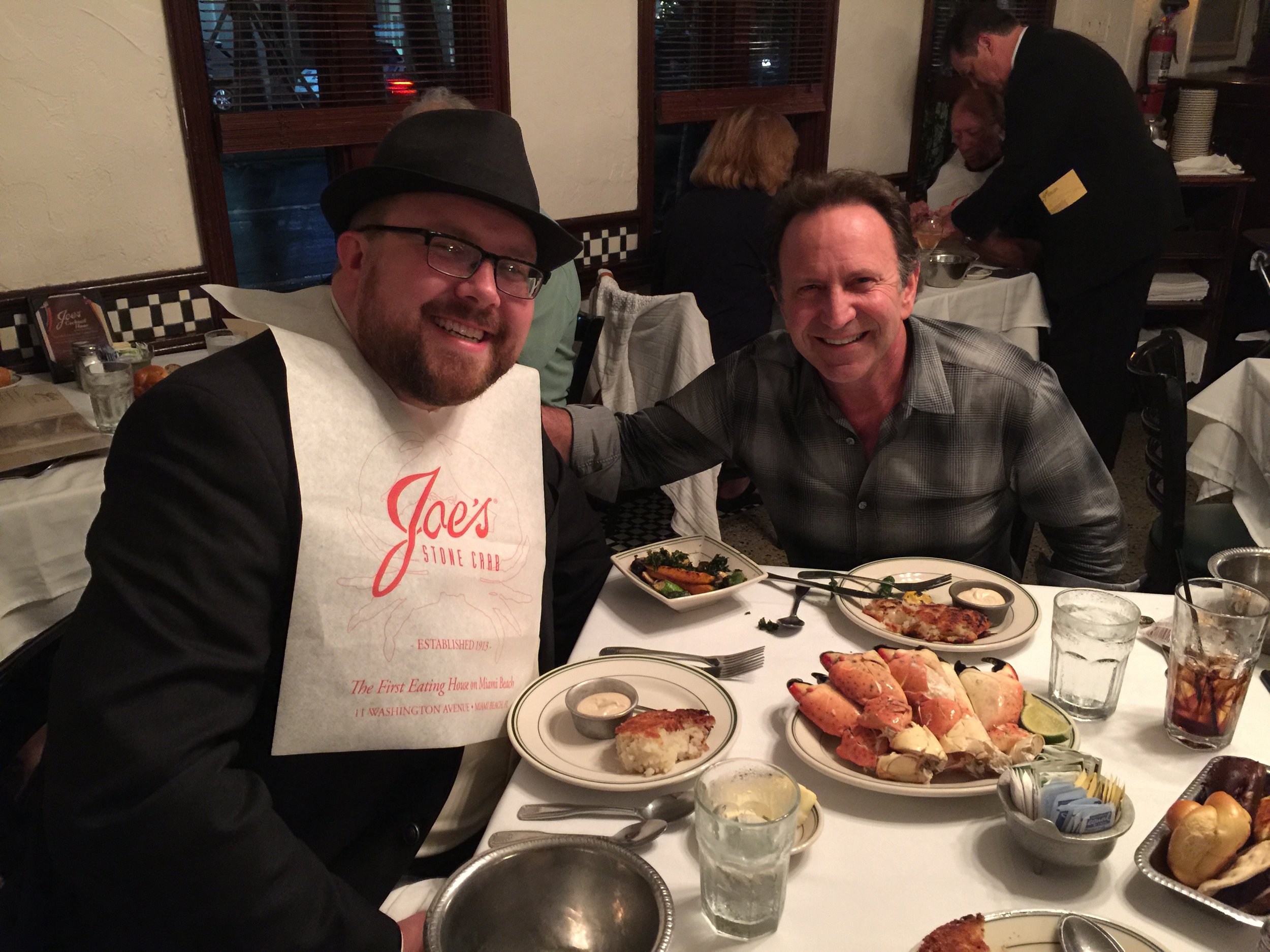 Two men smiling at a restaurant table with seafood dishes, drinks, and a bib reading 'Joe's Stone Crab'.