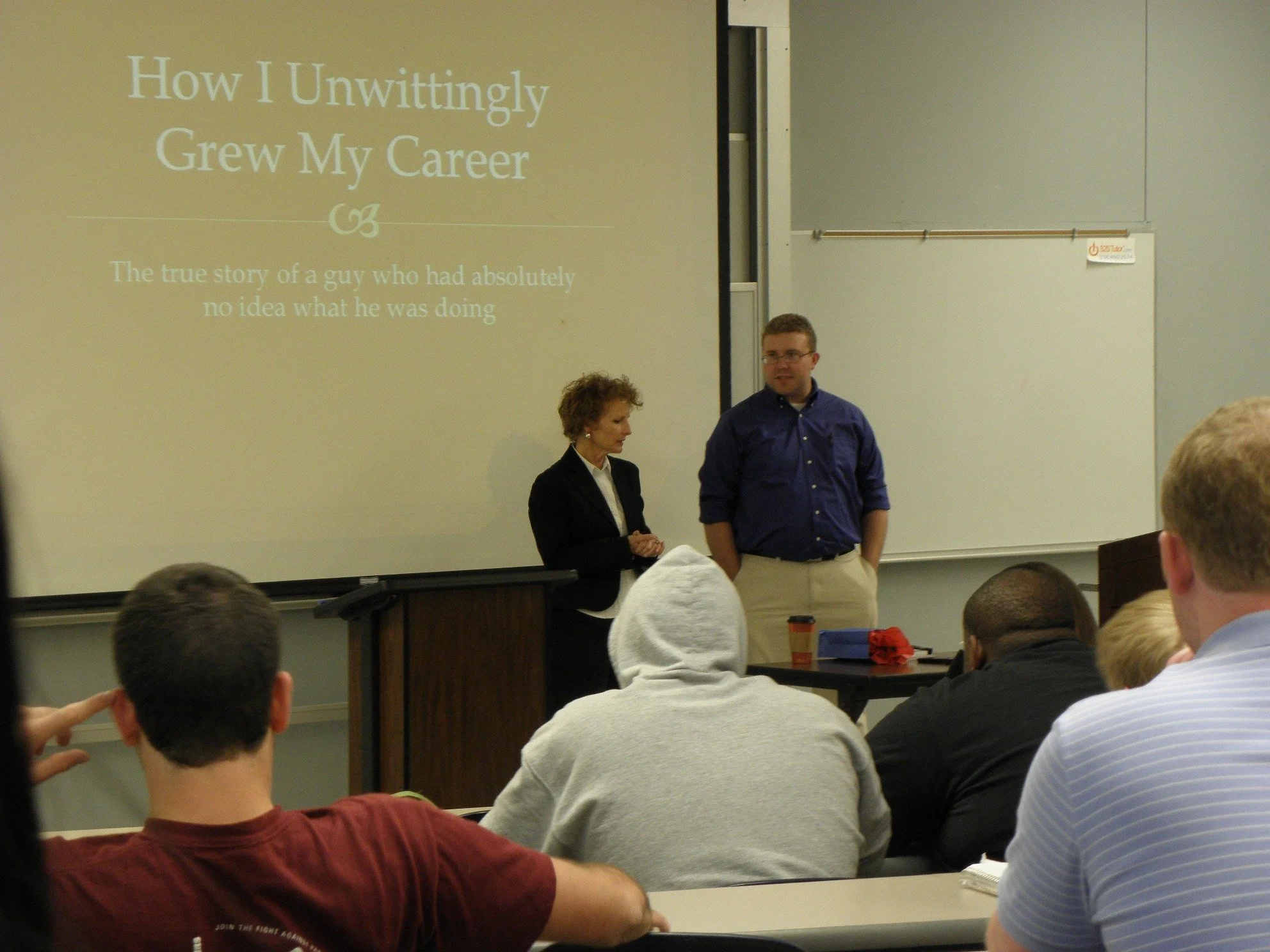 A classroom presentation titled 'How I Unwittingly Grew My Career' with two speakers at the front. Audience seated, with some taking notes, in a lecture hall with a whiteboard and projection screen.