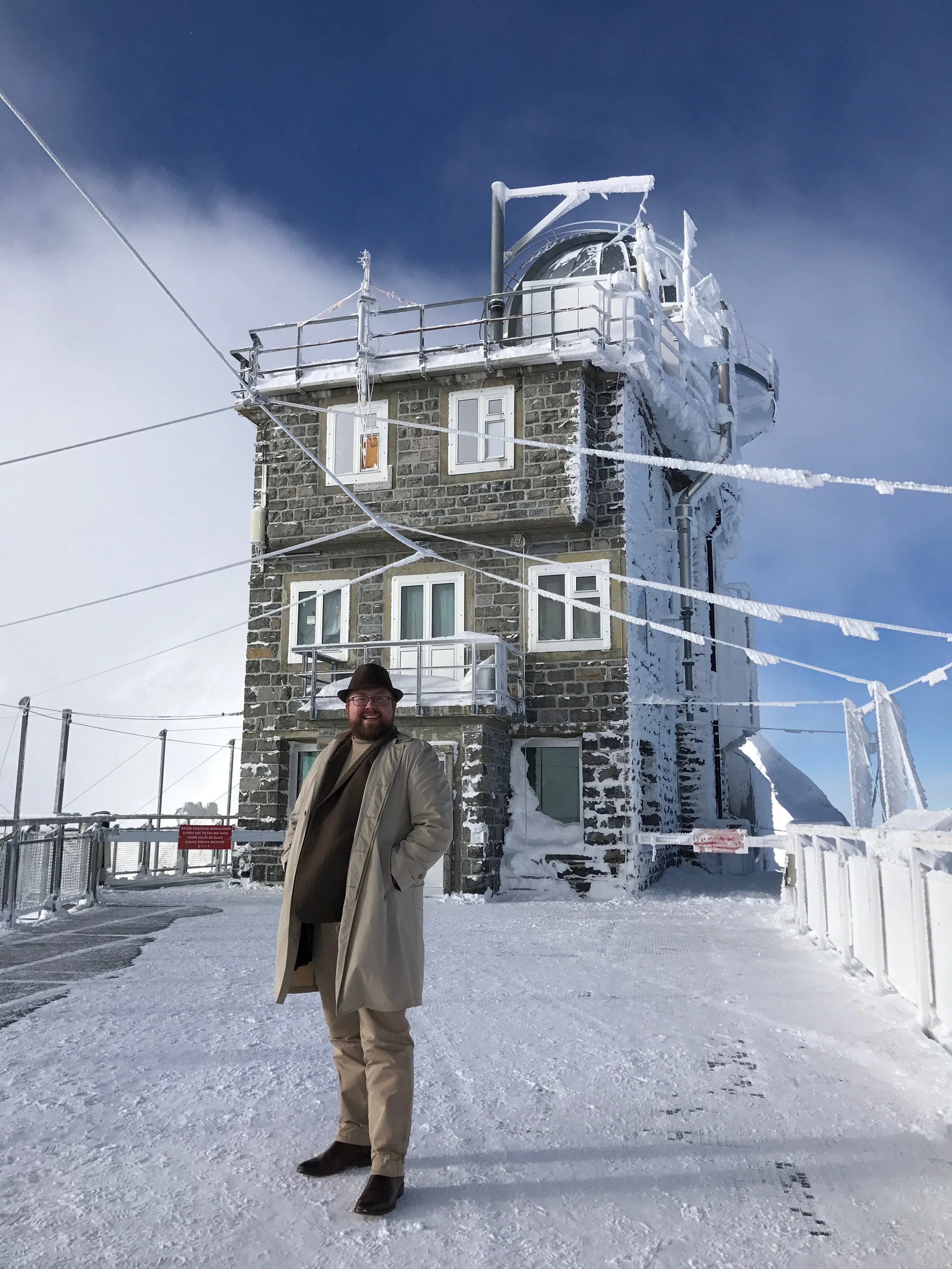 A man dressed in a beige coat, black hat, and glasses stands on snow-covered ground in front of a stone building with multiple windows and ice-covered railings. The container at the top of the building is also covered in snow and ice, with a clear bl