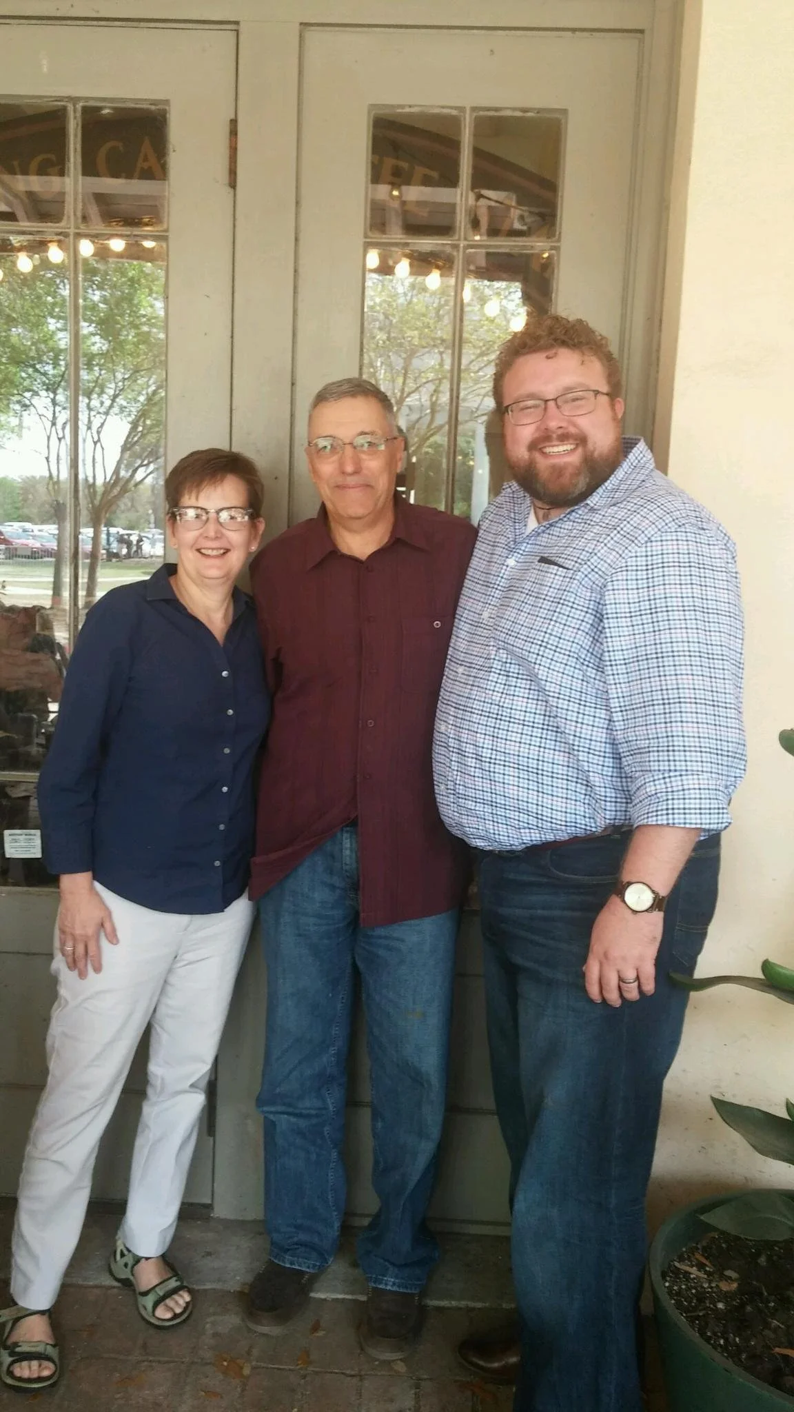 Three people standing outside near a glass door, smiling at the camera. A woman wearing a navy blue shirt and white pants on the left, a man in a burgundy shirt and jeans in the middle, and a man in a plaid shirt and dark jeans on the right.
