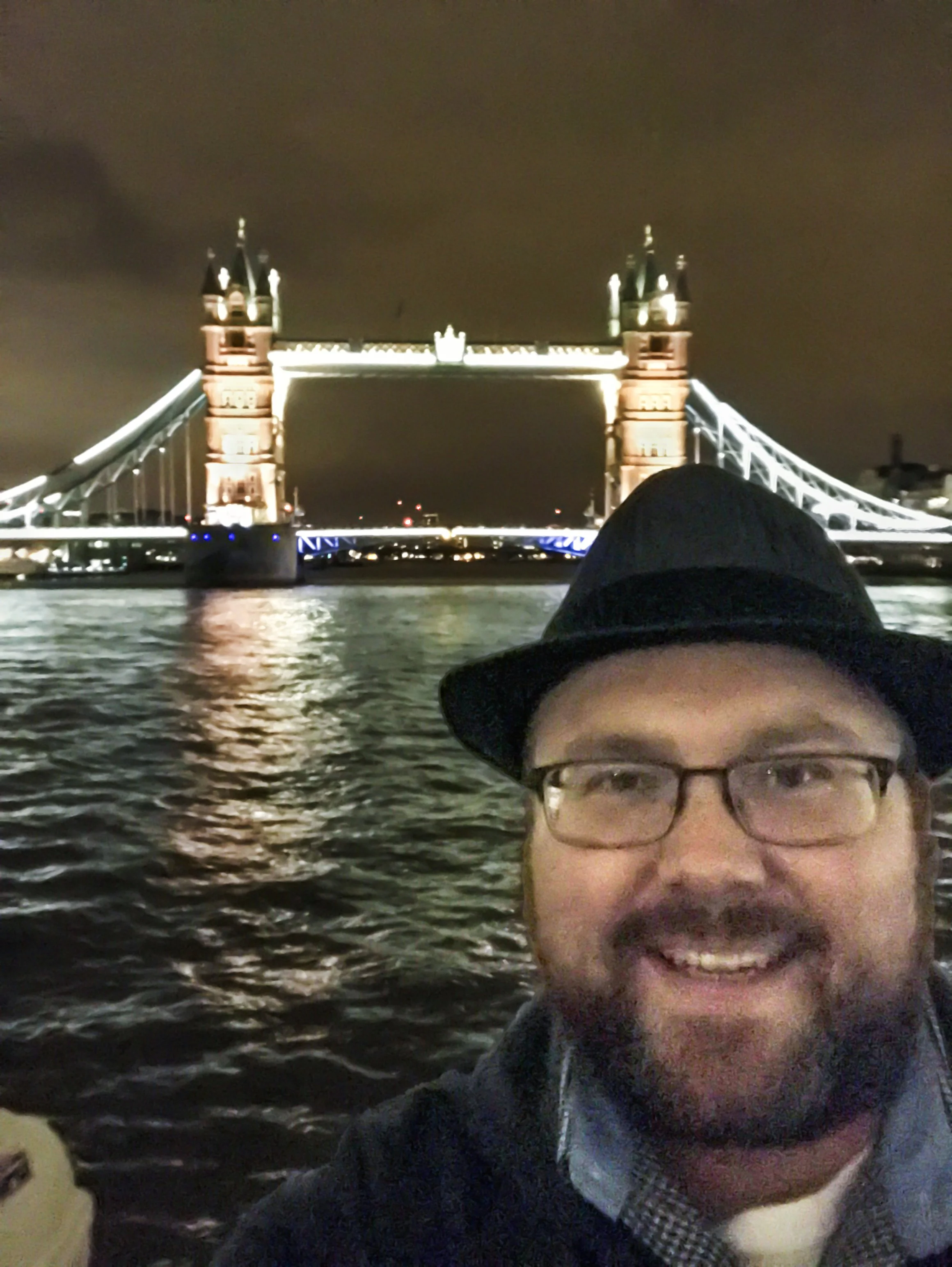 Selfie of a man wearing glasses, a fedora, and a dark jacket, smiling at night with Tower Bridge illuminated in the background over the River Thames.