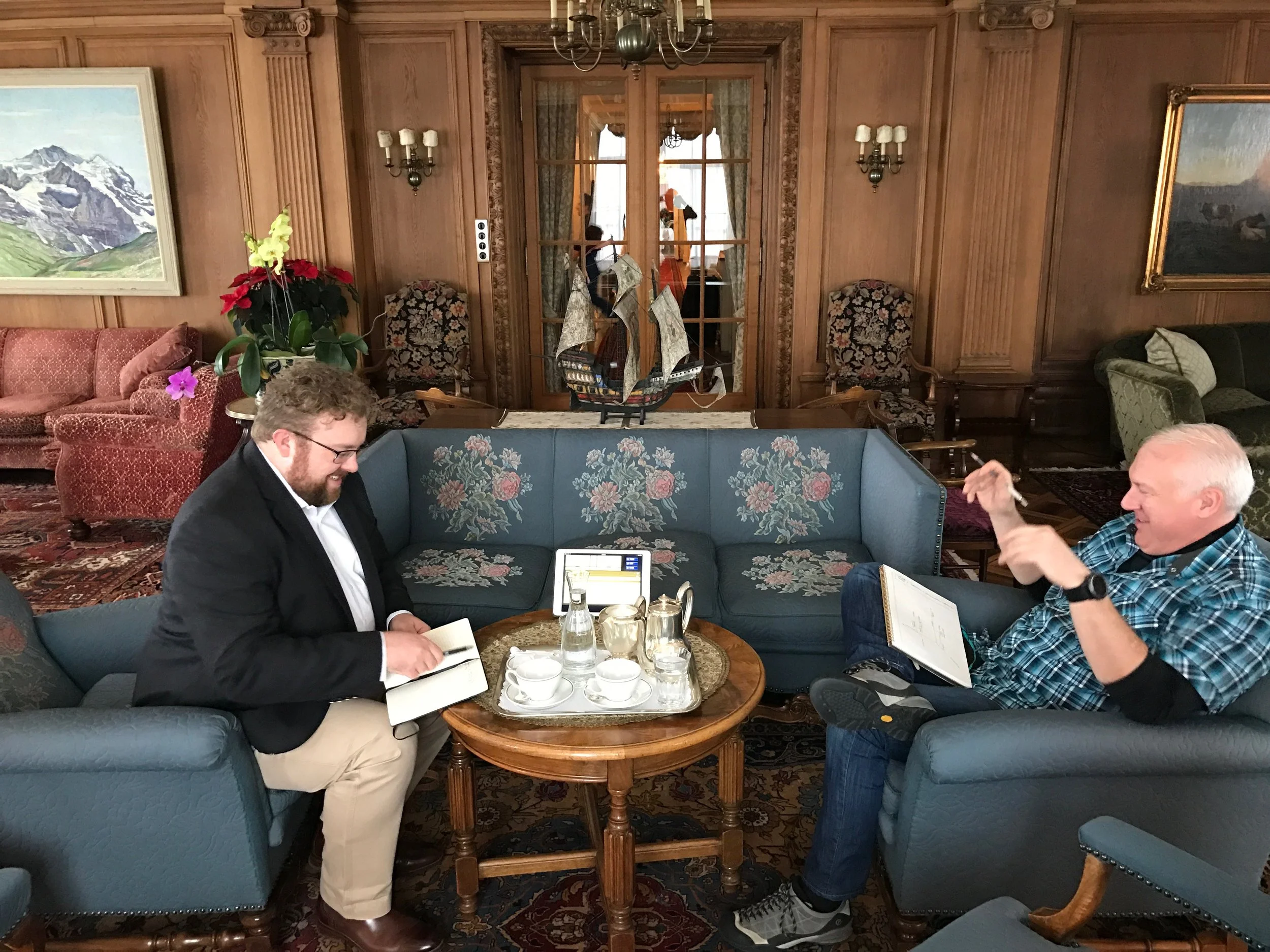 Two men sitting on blue armchairs around a small wooden table with teacups, a teapot, and water bottles in a wood-paneled lounge. One man is smiling and looking at a notebook; the other is gesturing with his hands and smiling.