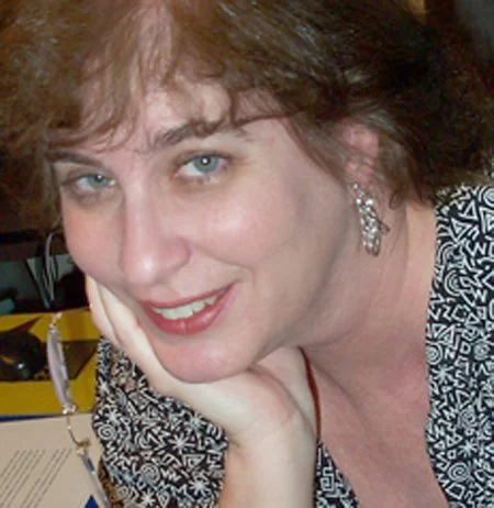 A woman with light skin, short curly reddish-brown hair, and blue eyes, smiling and resting her chin on her hand, wearing earrings and a patterned black and white top.