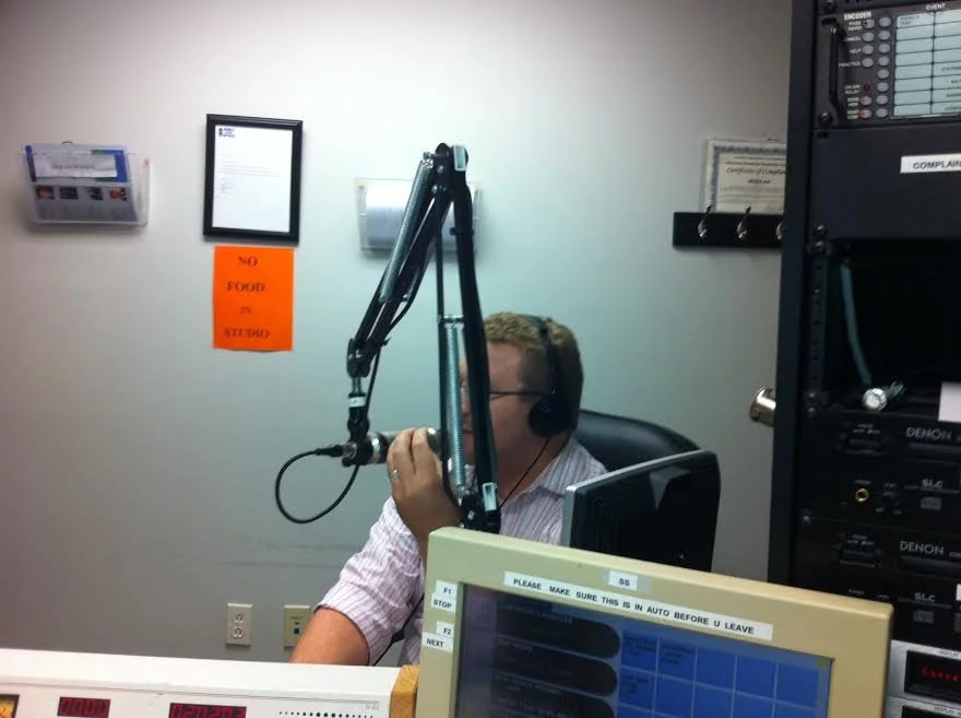 Radio host or DJ wearing headphones speaking into a microphone at a broadcasting station with audio equipment and monitors.