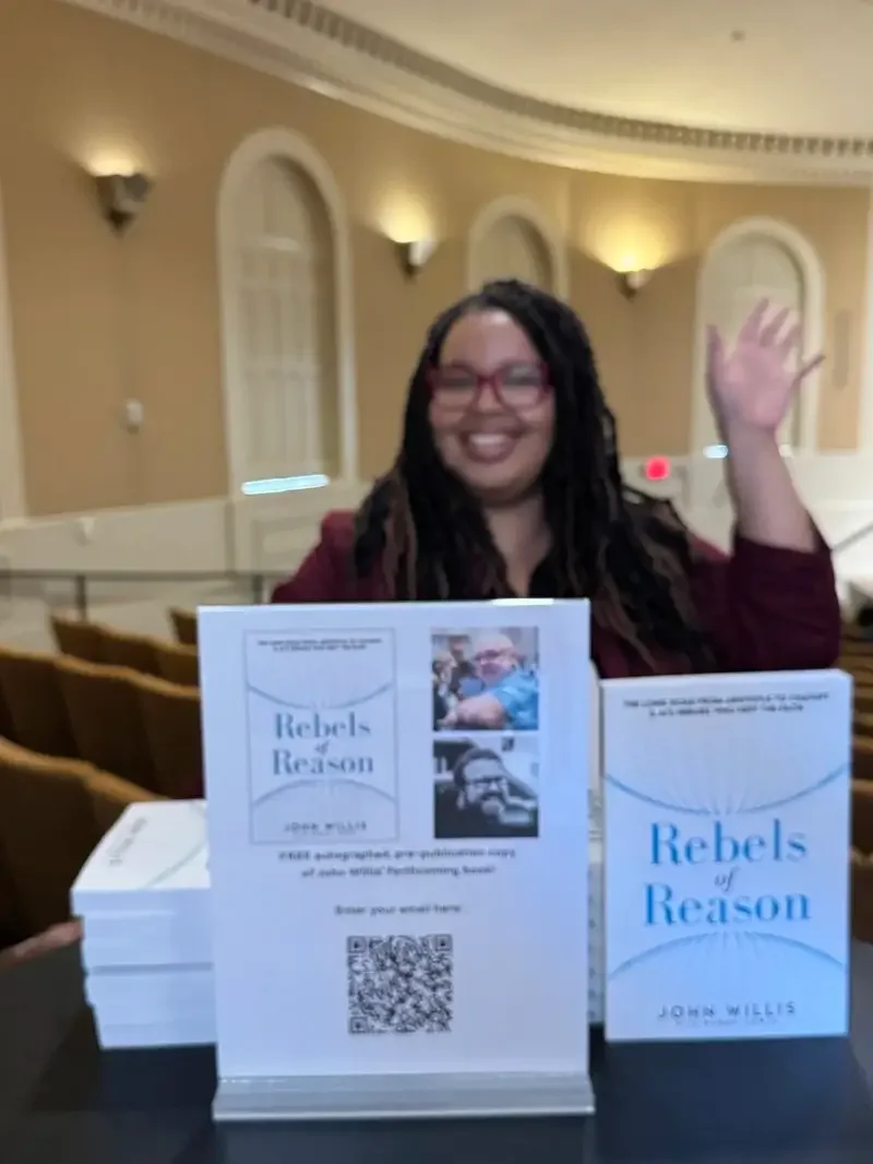 A woman with dreadlocks, glasses, and a maroon blazer sitting at a table, waving, with a stack of copies of the book "Rebels of Reason" by John Willis on the table in front of her.