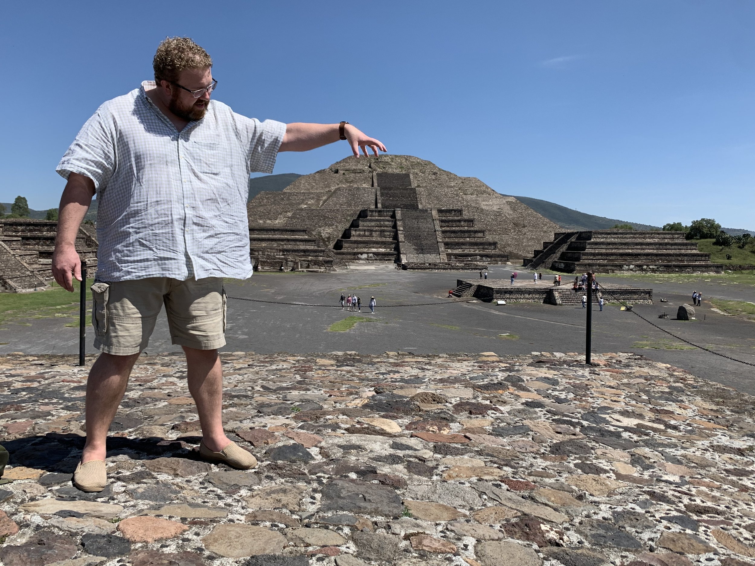 Man in shorts and glasses pointing at the Pyramid of the Magician at Uxmal archaeological site in Mexico on a clear day.