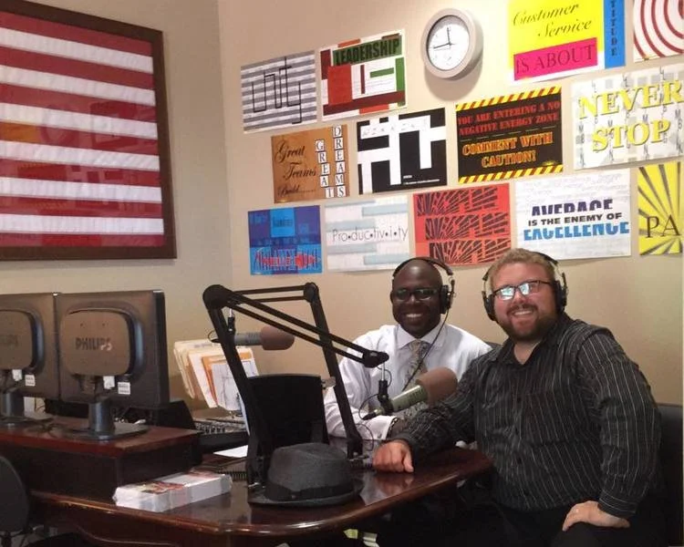 Two men sit at a radio station desk with microphones and headphones, smiling at the camera. The wall behind them is decorated with colorful signs and motivational posters. A large American flag is displayed on the left.