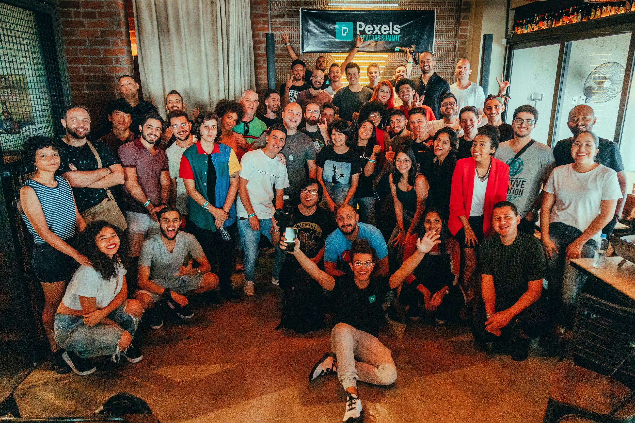 A large group of diverse people at an indoor event, smiling and posing for a group photo in front of a banner that reads 'Pexels Creators Summit'.