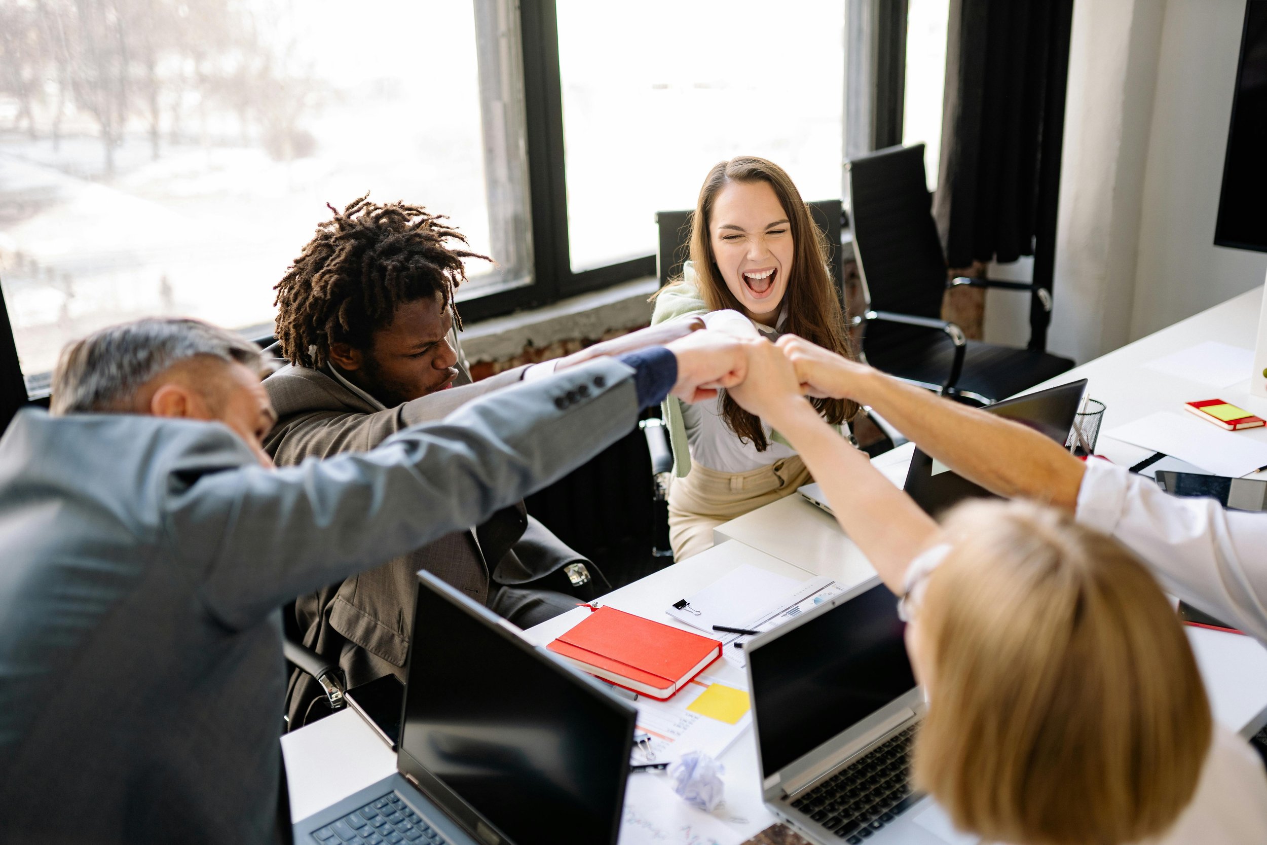 Group of diverse coworkers in a meeting room, united in a fist bump and smiling, with laptops, notebooks, and papers on the table.