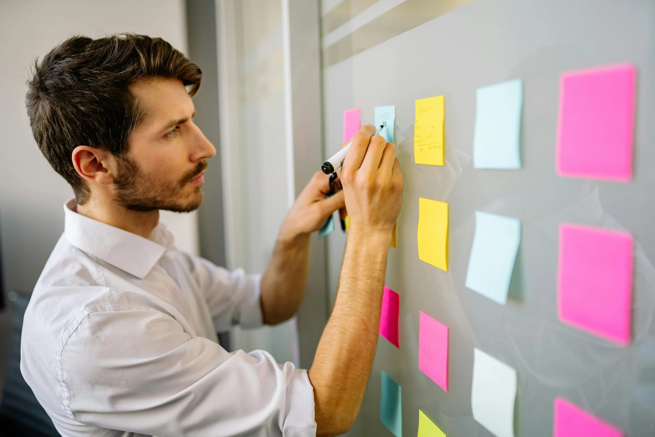 A man in a white shirt writing on colored sticky notes placed on a whiteboard, with notes in pink, blue, and yellow.