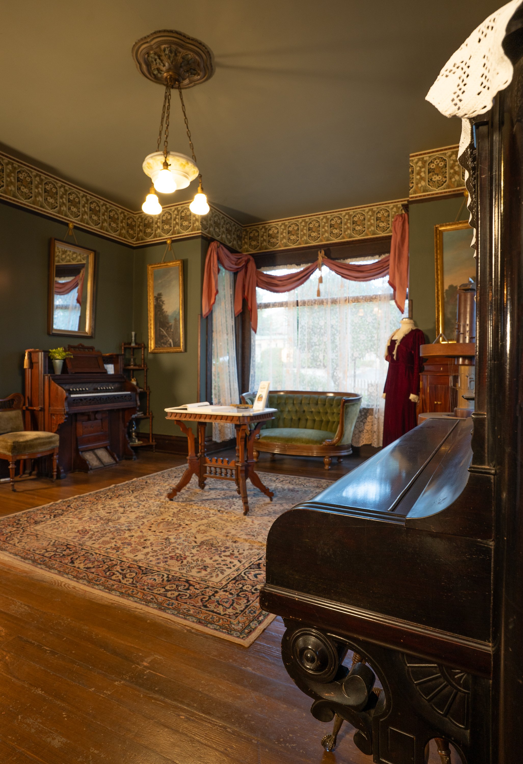 A vintage living room featuring a dark wooden piano, a green velvet sofa, antique framed landscape paintings, a patterned area rug, a chandelier with three light bulbs, ornate curtains with valances over large windows, and a mannequin dressed in a red vintage dress.