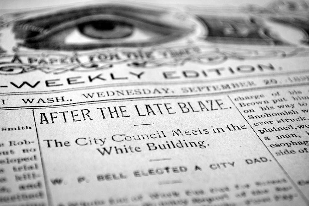 Close-up of a black and white newspaper with a prominent eye illustration at the top and headlines, including 'After the Late Blaze' and 'The City Council Meets in the White Building'.
