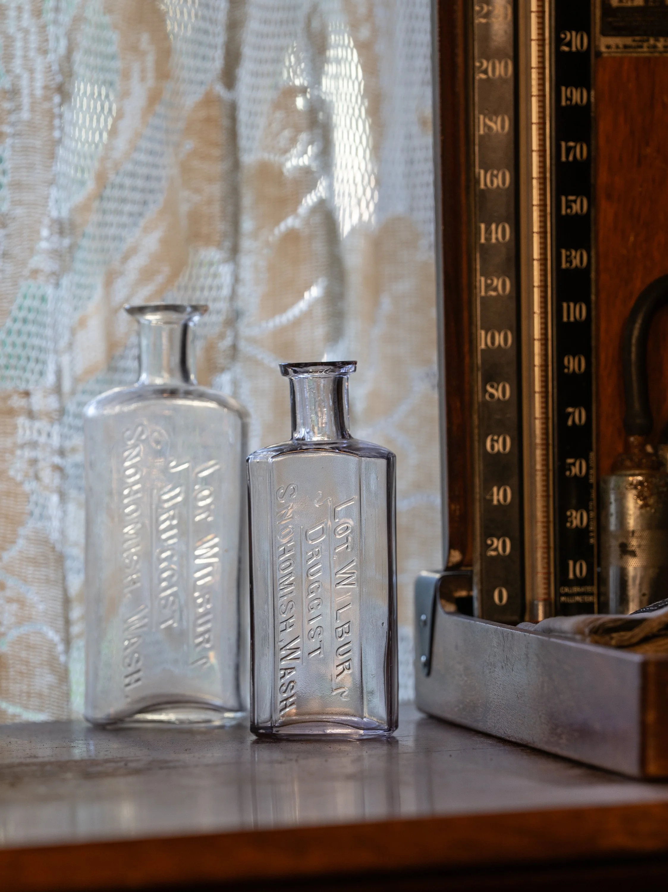 Close-up of vintage glass bottles on a wooden shelf, with a cloudy window curtain in the background, and a vintage barometer with black and gold markings to the right.