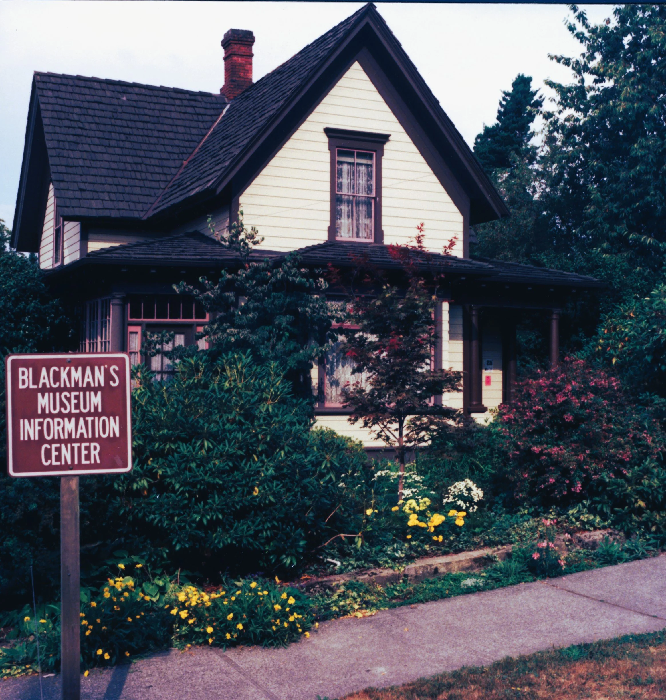 A house with a steep gabled roof, cream-colored siding, a chimney, and a front window. There is a garden with colorful flowers in front and a street sidewalk.
