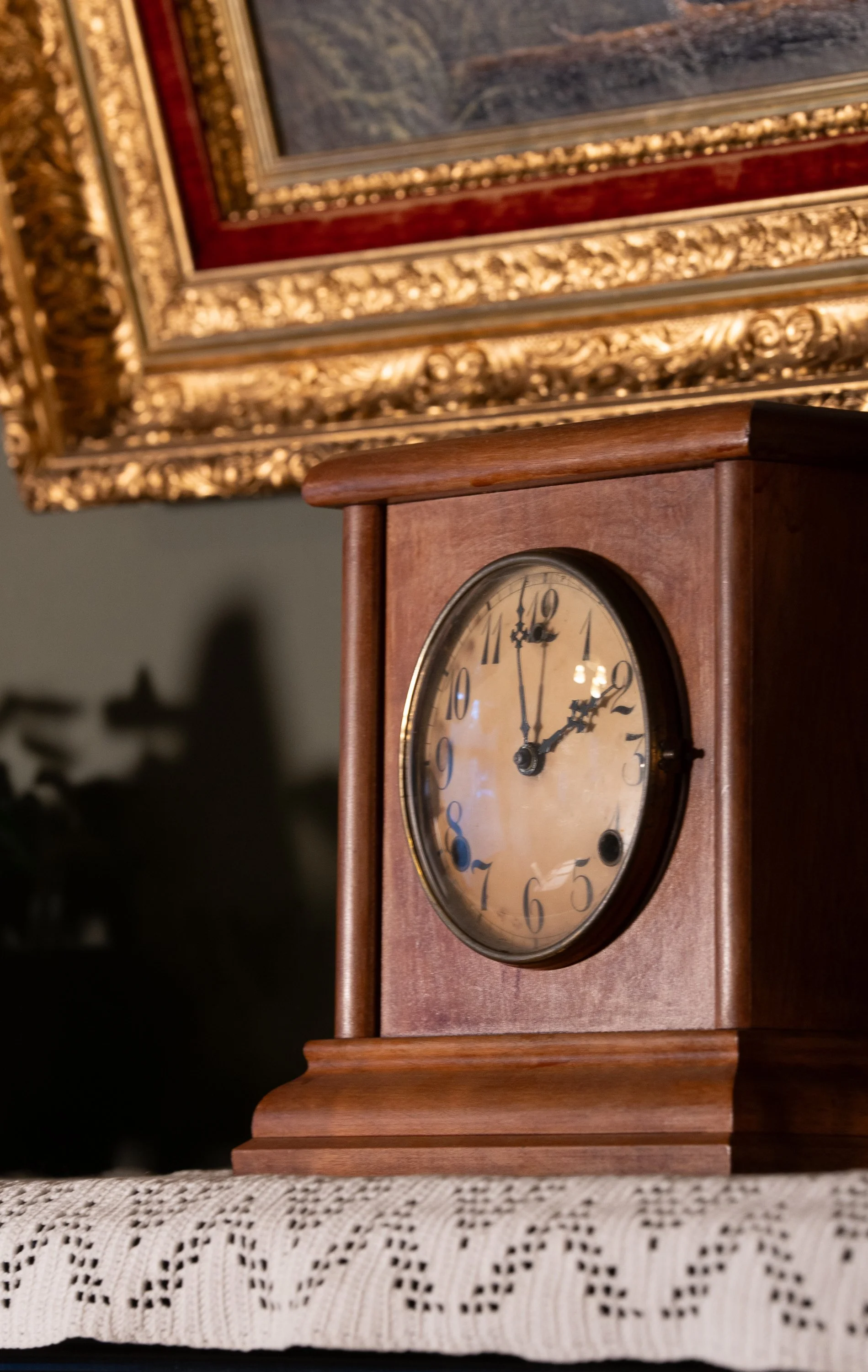 A vintage wooden clock with a round face showing the time 2:56, placed on a lace tablecloth with a framed painting with ornate gold and red details hanging on the wall in the background.