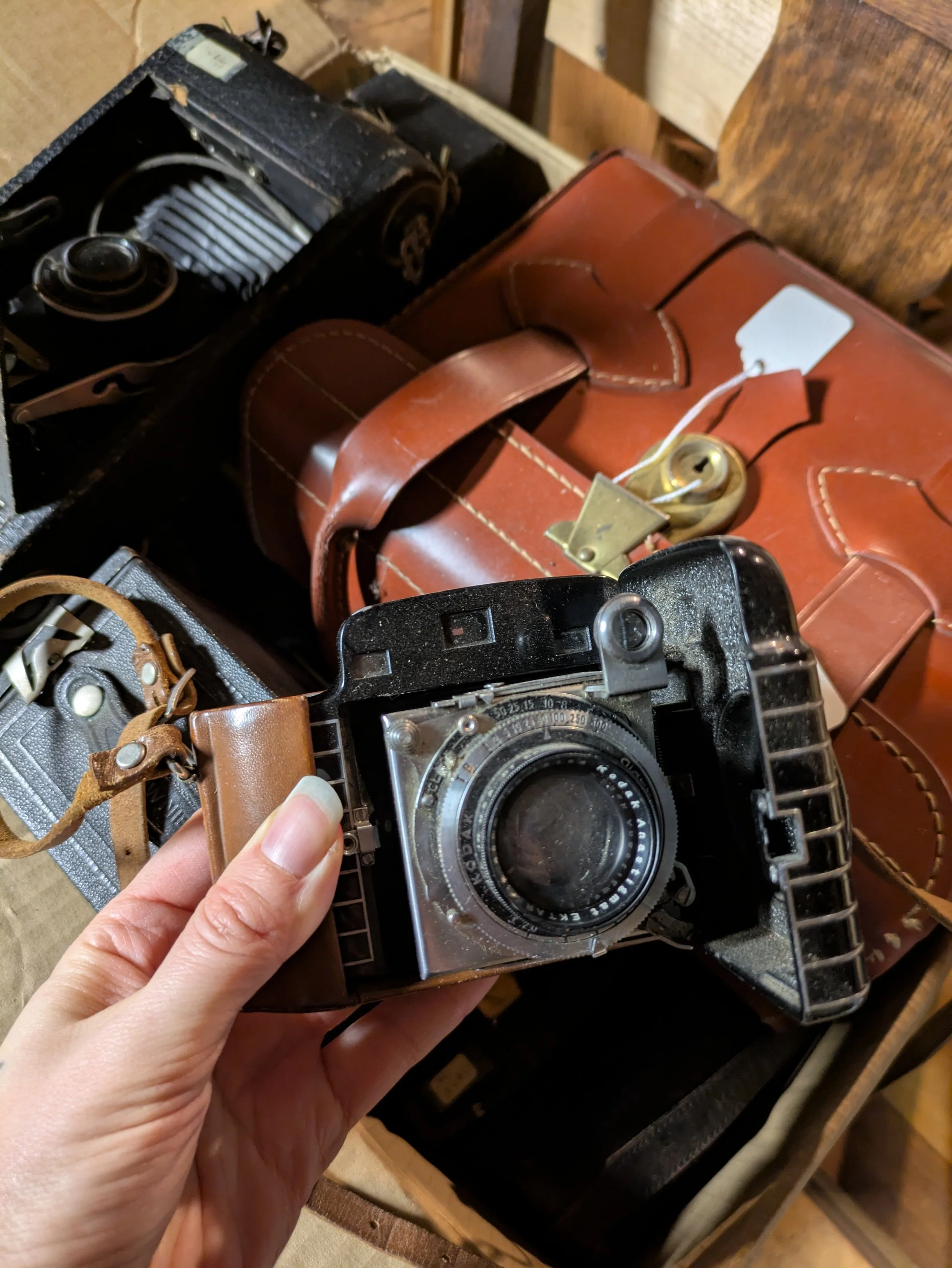 A person holding a vintage camera with a leather strap, with a brown leather bag and other vintage items on a wooden surface in the background.