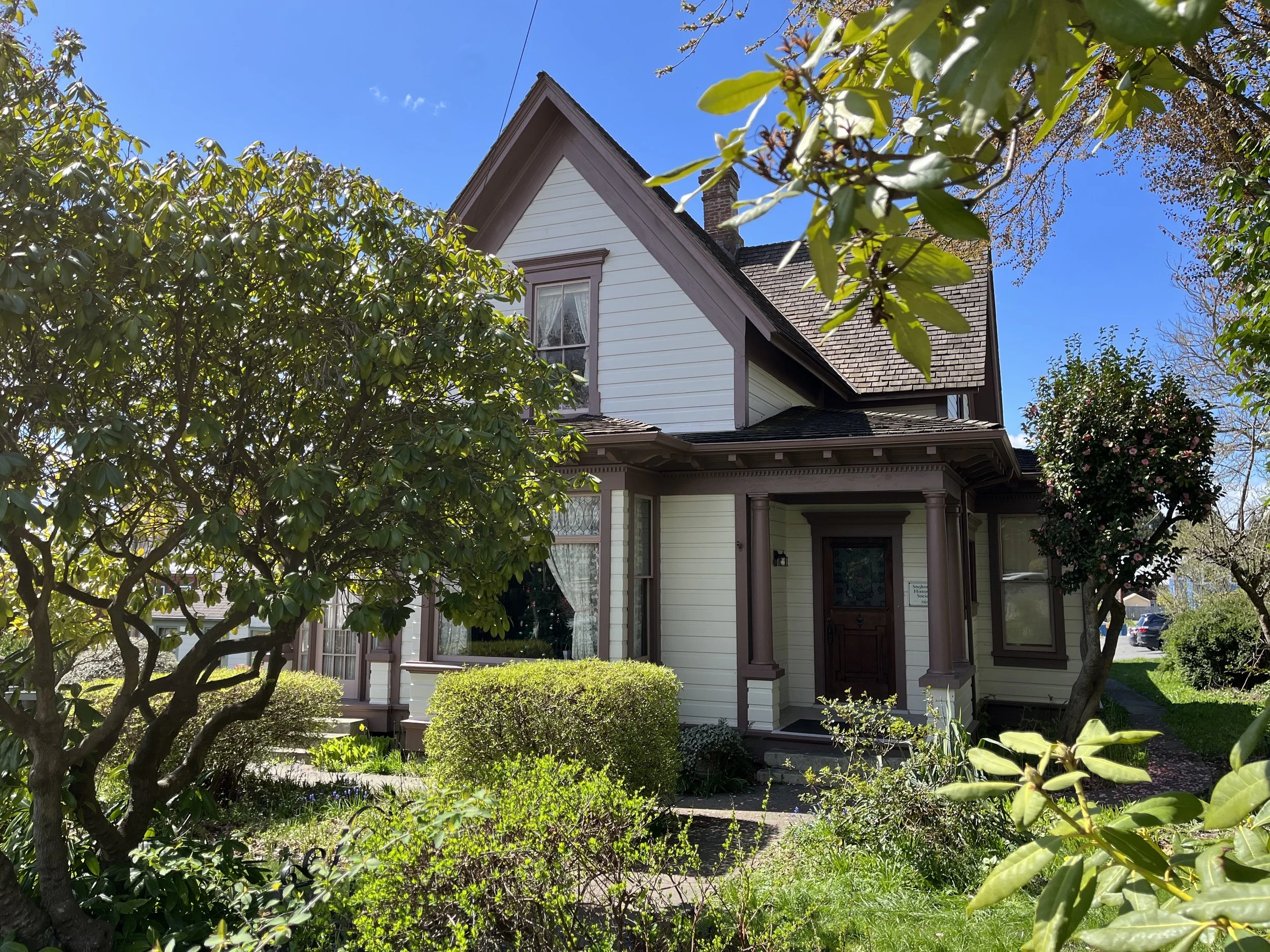 A house with a gable roof, white siding, and a front porch, surrounded by trees and a garden with bushes and flowers, under a clear blue sky.