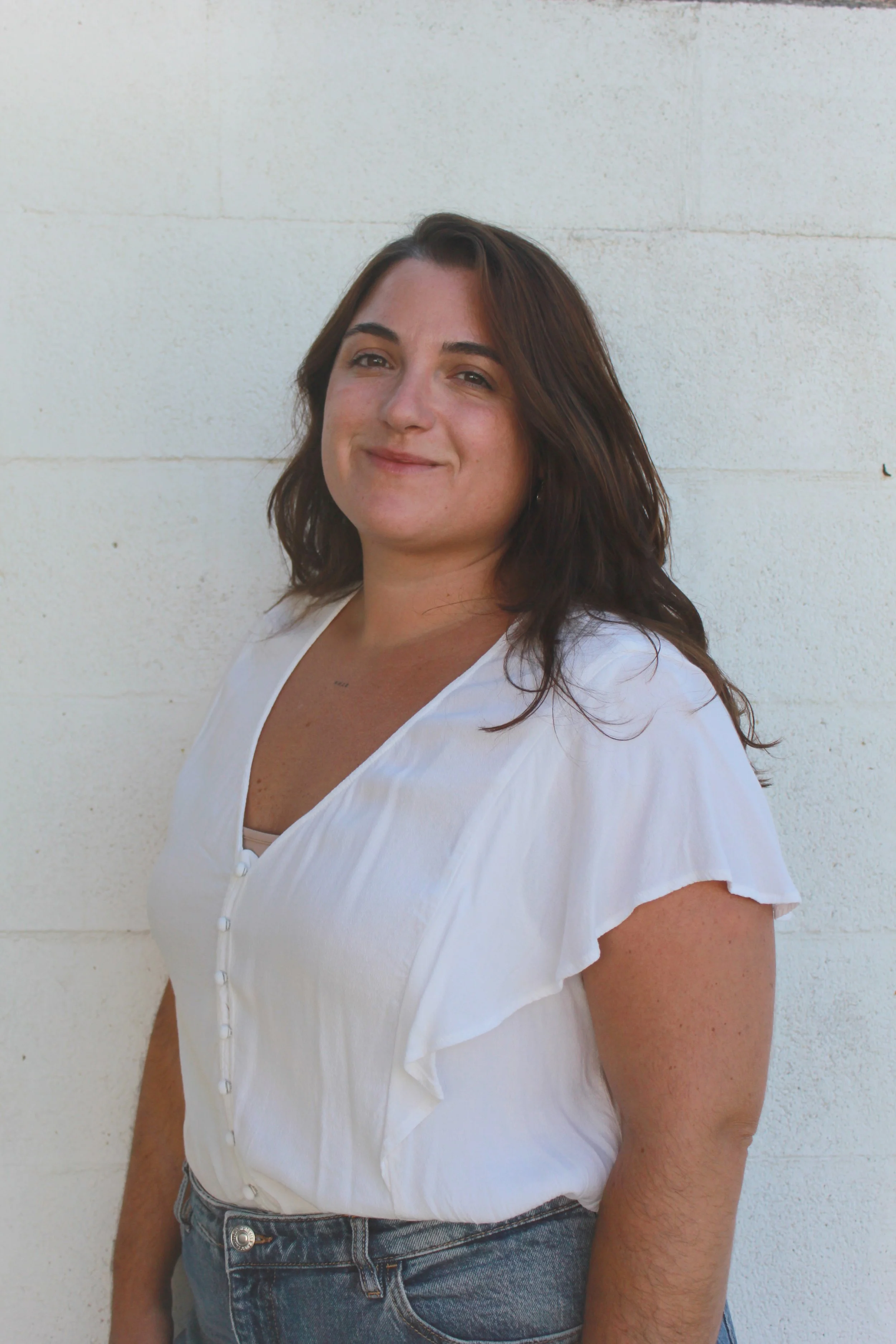 Mujer con cabello castaño oscuro, usando blusa blanca con botones y jeans, sonriendo ligeramente, de pie contra una pared blanca.