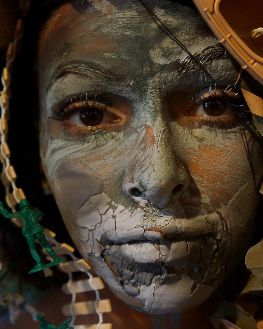 📷 La Femme! Framing the Feminine Exhibit
&ldquo;Rios Becomes Navy&rdquo; by Nancy Hines

A face transformed into battlefield and myth &mdash; cracked clay, plastic soldiers, flowers, and memory woven together, revealing the weight a woman carries be