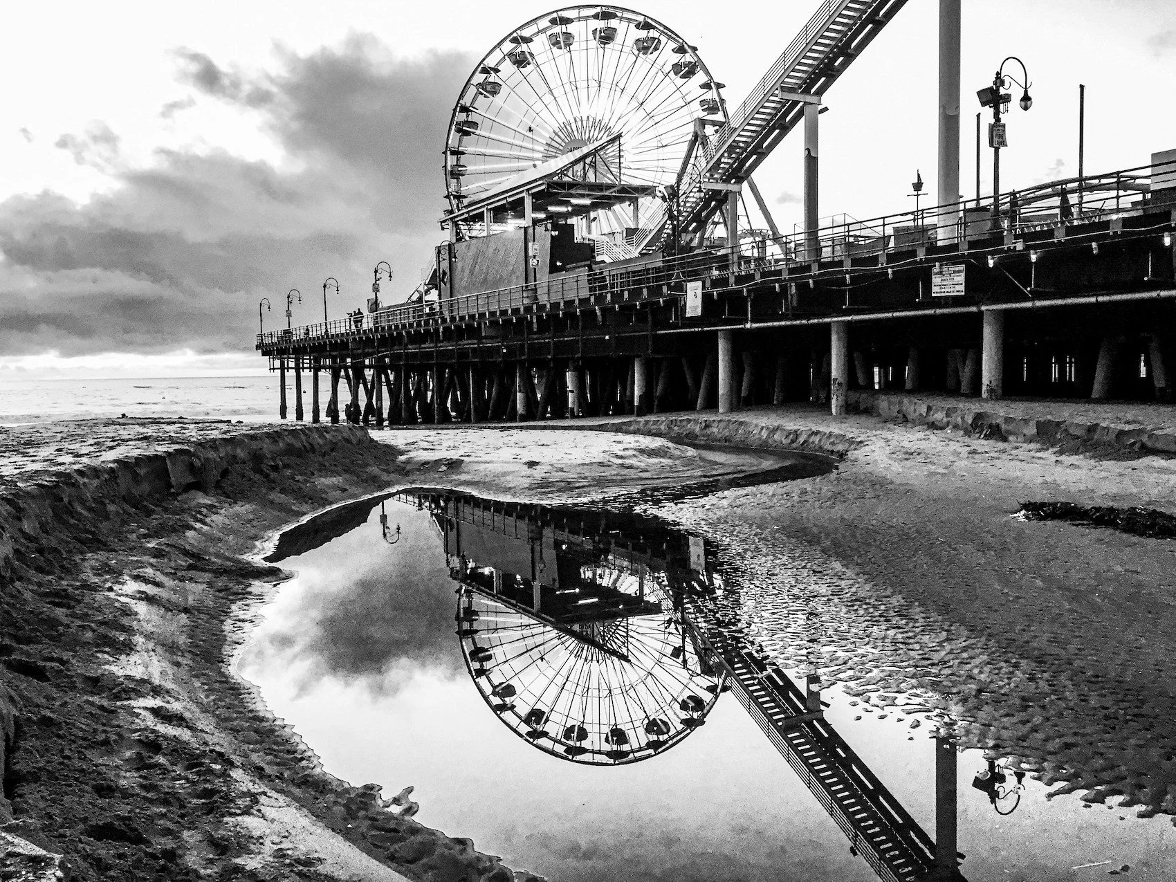 📷 B&amp;W: The Poetry of Light &amp; Shadow
&ldquo;Carousel and Reflection&rdquo; by Marilyn Brodwick (@marbrodwick)

The quiet geometry of the pier meets the stillness of a tide pool, where the Ferris wheel doubles itself in reflection. What is usu