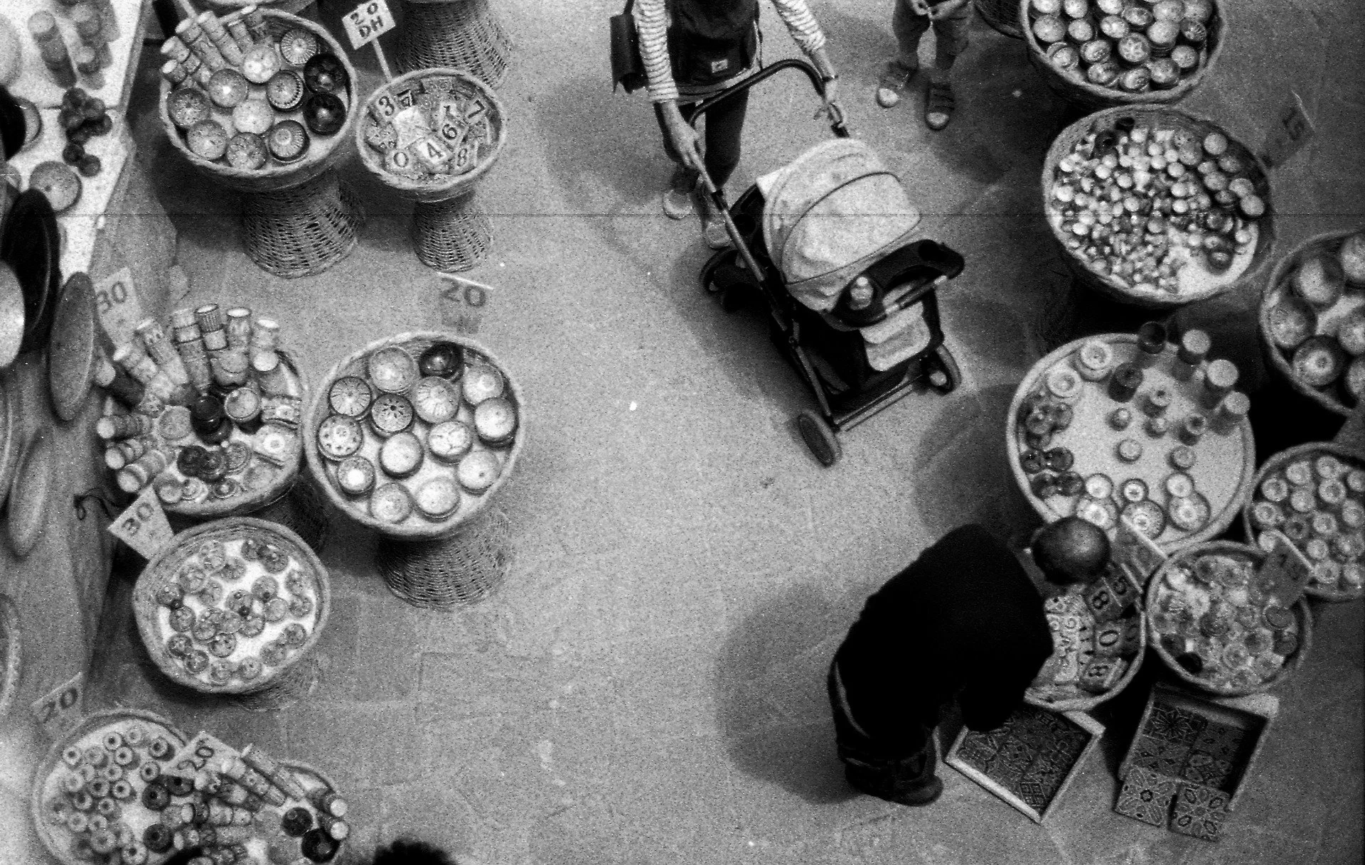 📷 B&amp;W: The Poetry of Light &amp; Shadow
&ldquo;Between Baskets&rdquo; by Louis Bryant

Seen from above, baskets of goods form a quiet rhythm around the small movements of daily life &mdash; a shopper, a stroller, a moment of pause within the pat