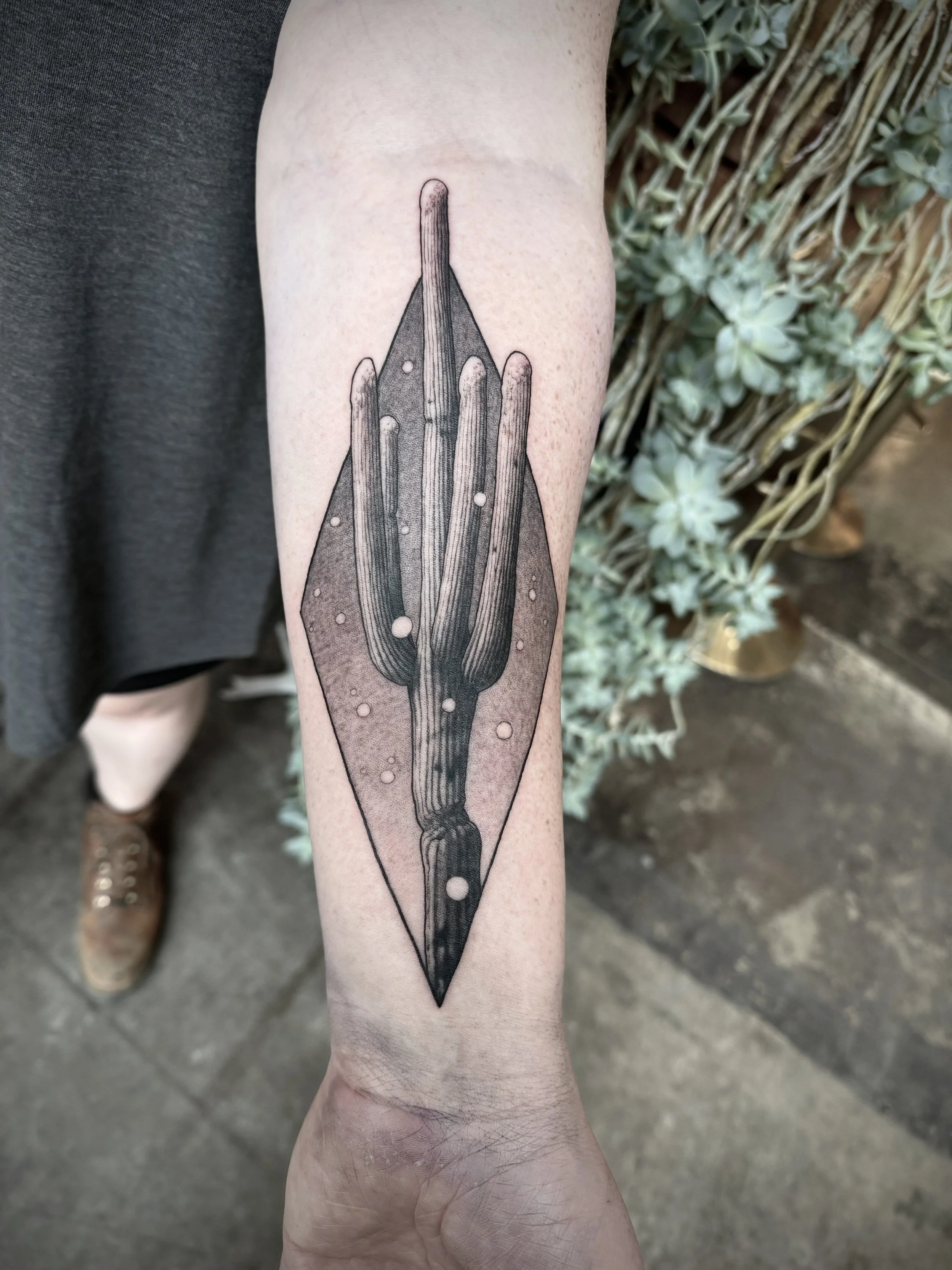 A black ink tattoo of a geometric dagger on a person's forearm, with a diamond-shaped outline surrounding the dagger. The background includes some green plants and a concrete floor.