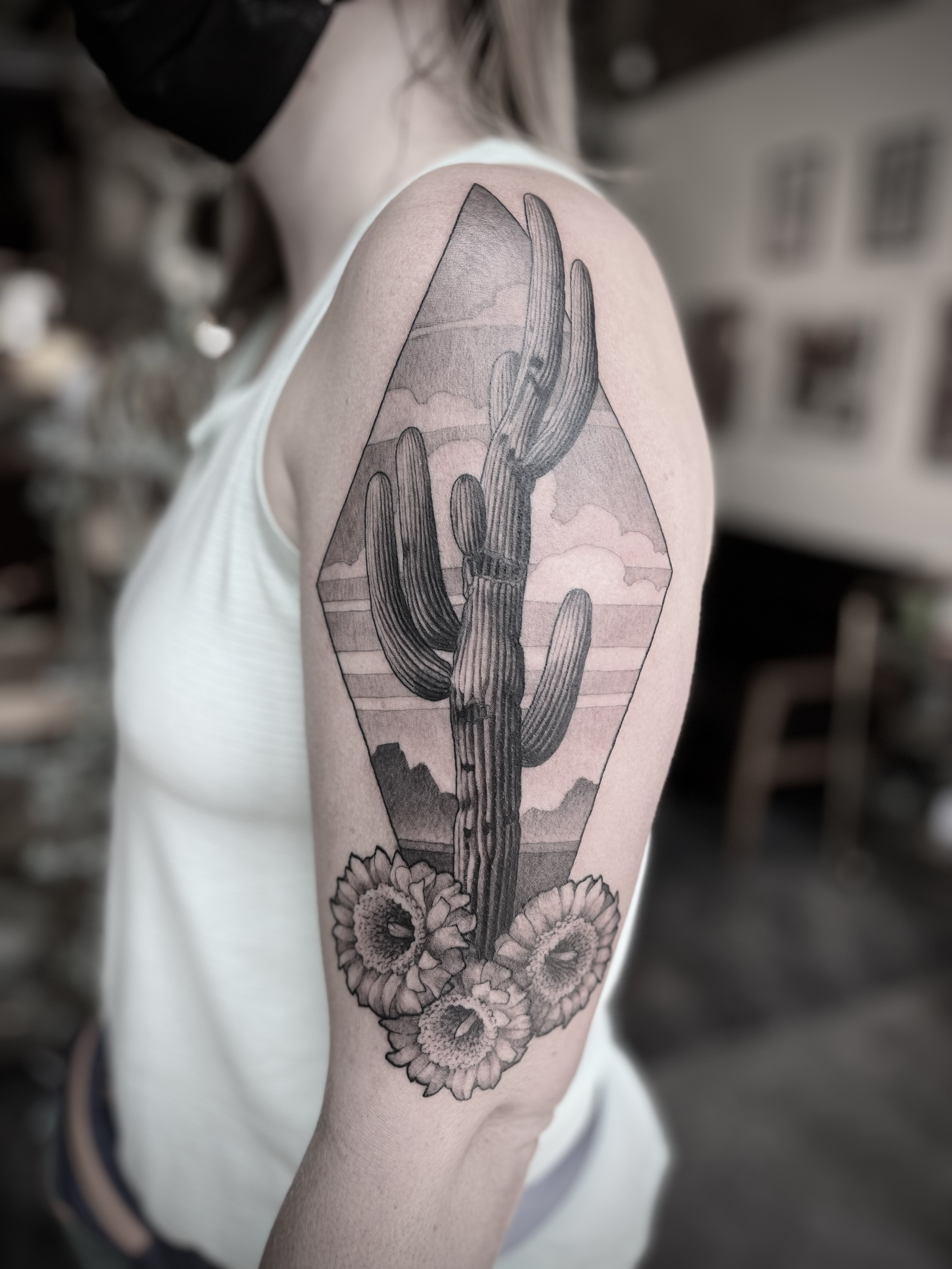 Black and gray tattoo of a desert landscape with cactus and mountains, surrounded by flowers, on a person's upper arm.