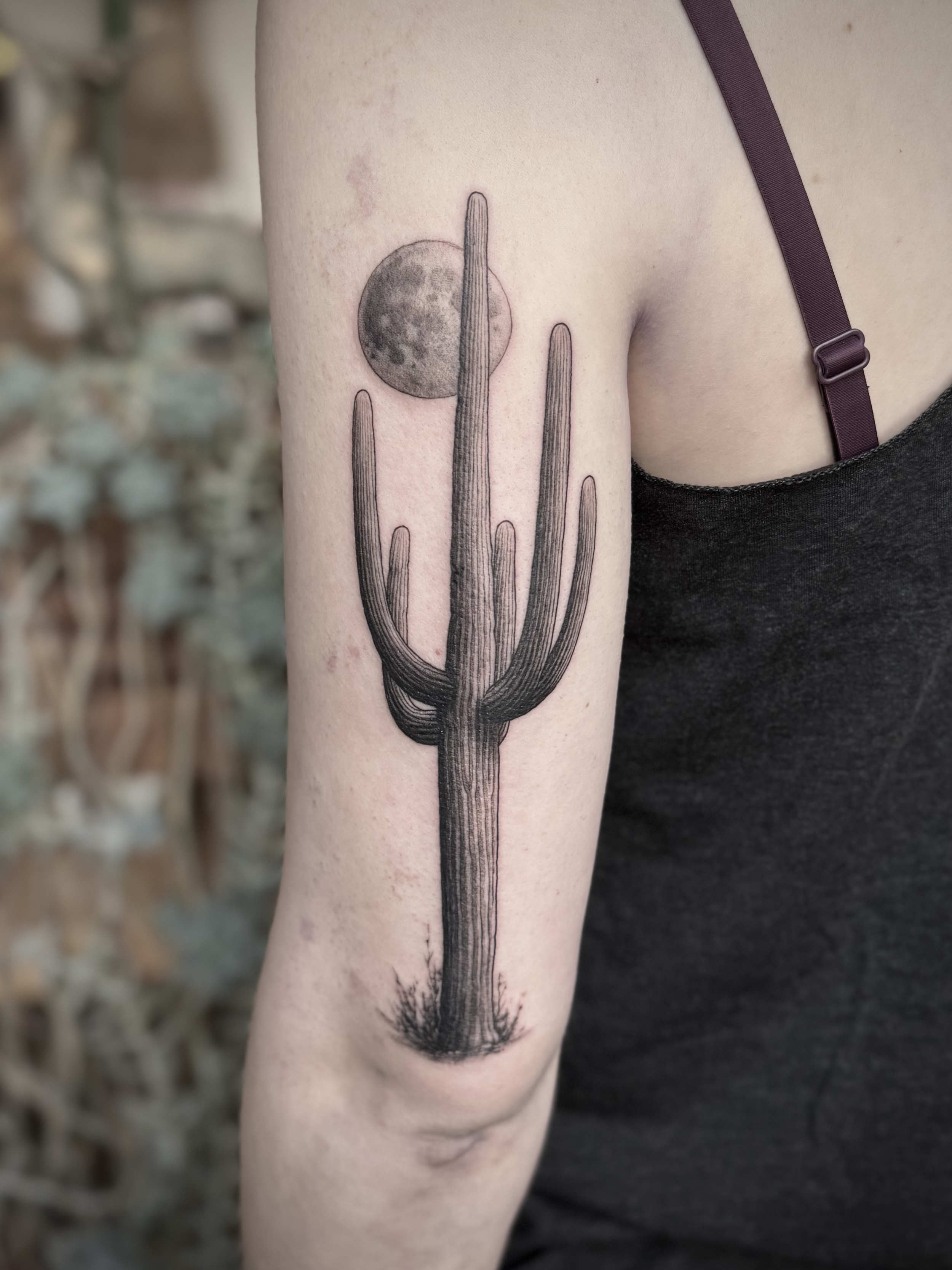 A black and white tattoo of a saguaro with some foliage at the base and a moon behind it. The tattoo is on the back upper arm of a woman.