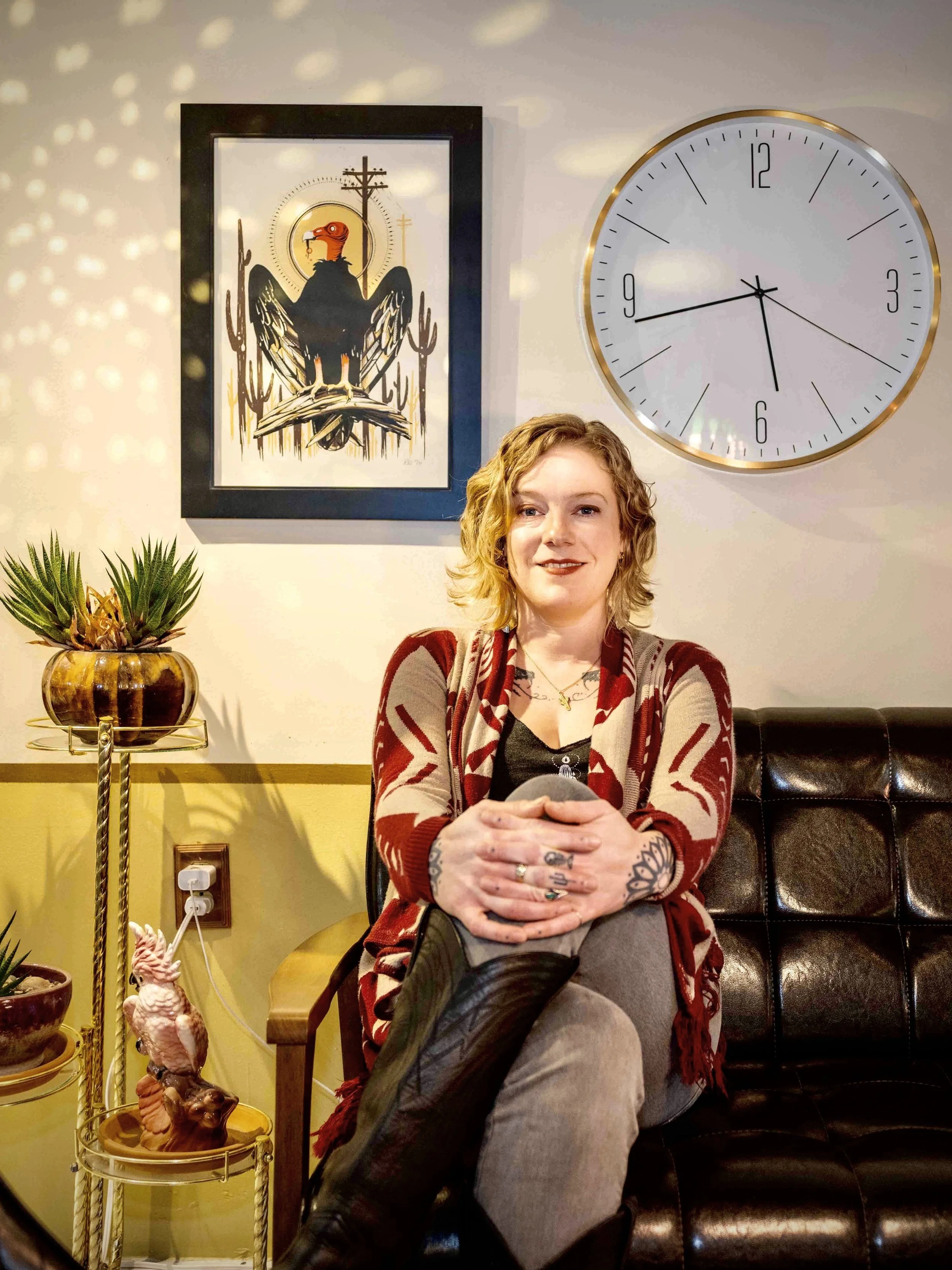 A woman with blonde hair sits in a brown leather chair, surrounded by  a large white clock and a framed design of a vulture. She is dressed in a red and white patterned cardigan over a black top.