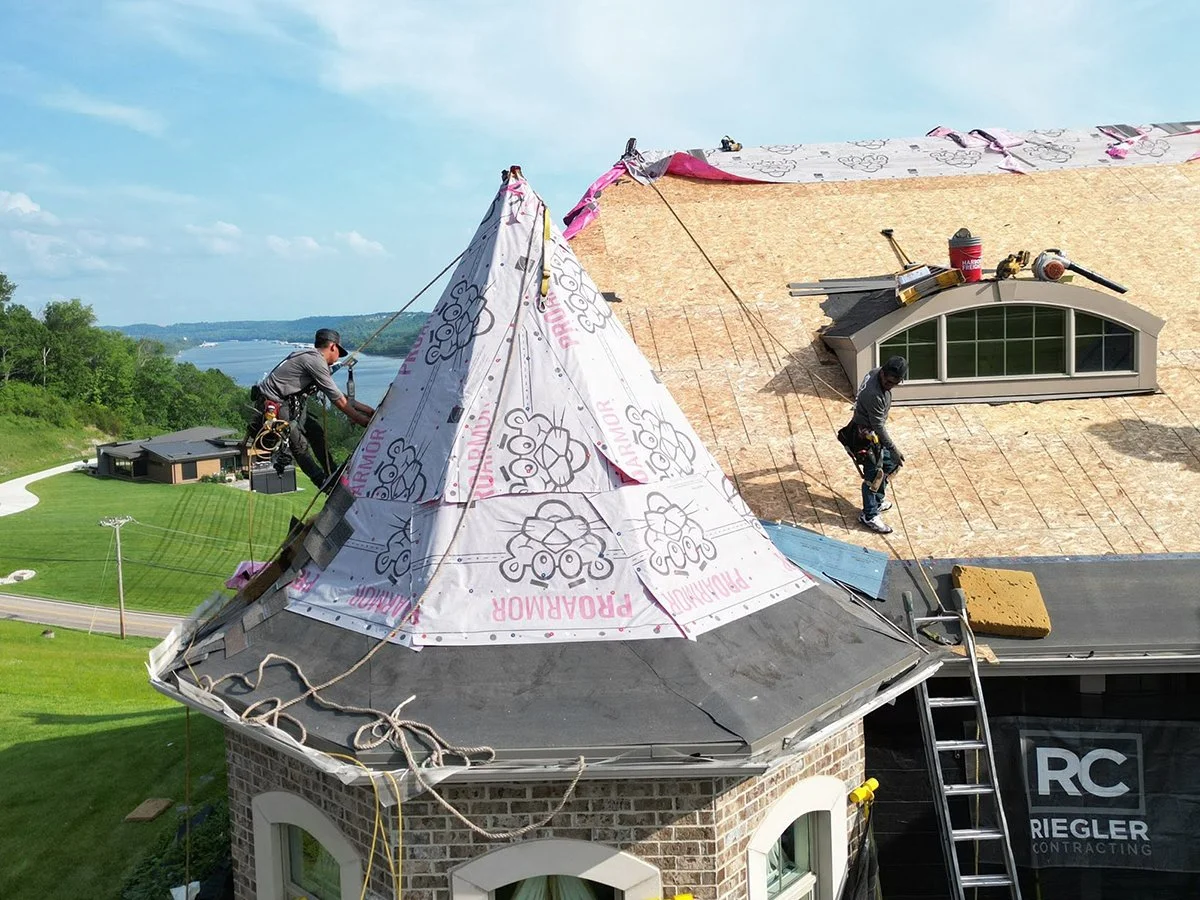 A photo of roofers replacing a roof in Cininnati, Ohio with the Ohio River in the background and a tarp protecting the home's landscaping with a Riegler Contracting logo on it.