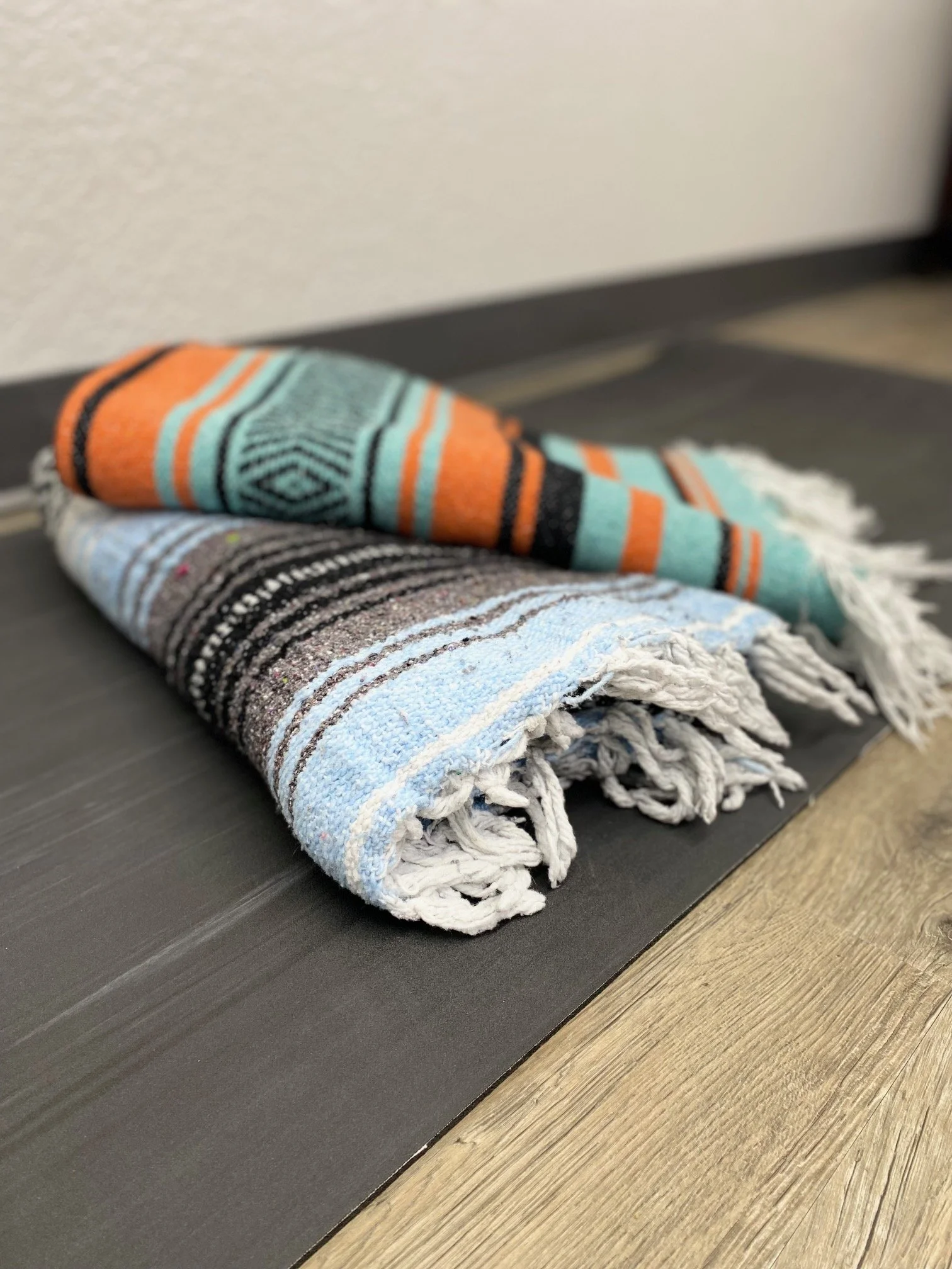 Two folded colorful striped yoga blankets on a black mat on the floor near a wall.