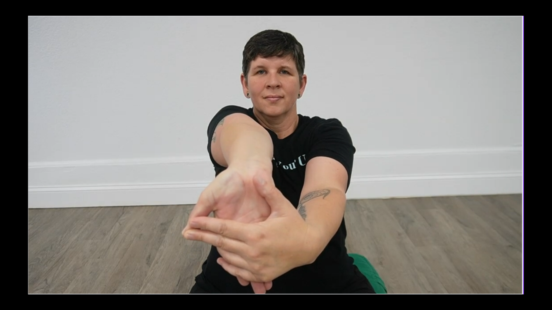 Woman with short hair and tattoos on her arms, sitting on the floor, stretching one arm forward and holding her fingers with the other hand, in a room with light-colored walls and wood flooring.