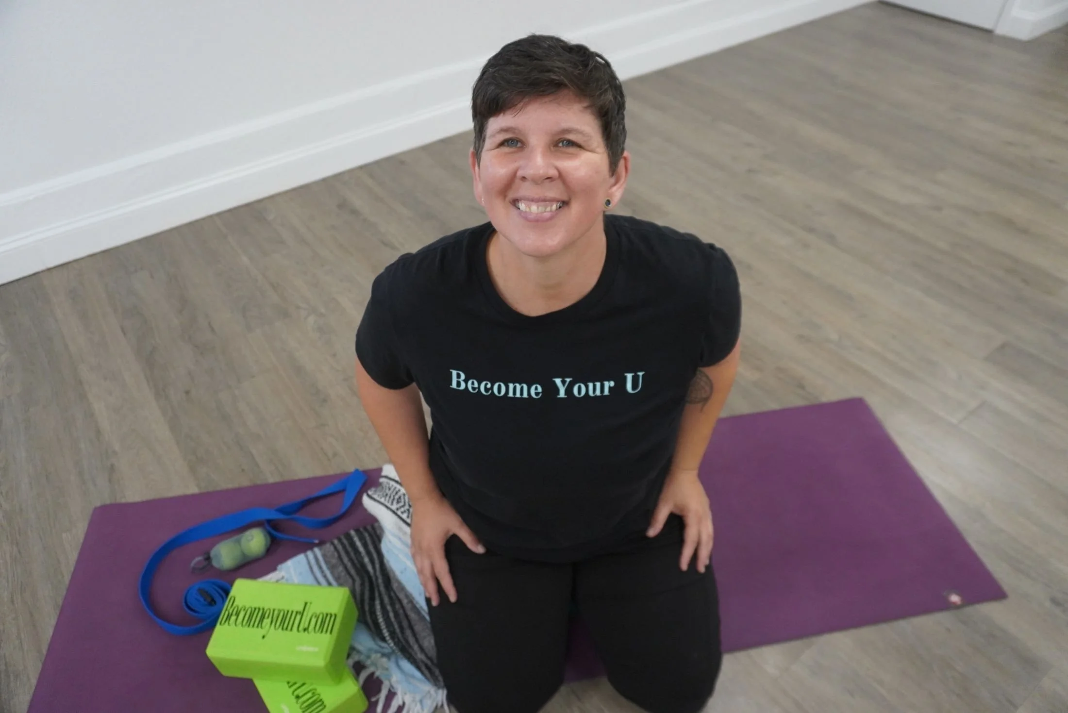 A smiling woman with short dark hair kneeling on a purple yoga mat indoors, wearing a black T-shirt with the words 'Become Your U' printed on it, surrounded by fitness items including a green jump rope, green and gray hand weights, and a green yoga block, in a room with light-colored wooden flooring and white walls.