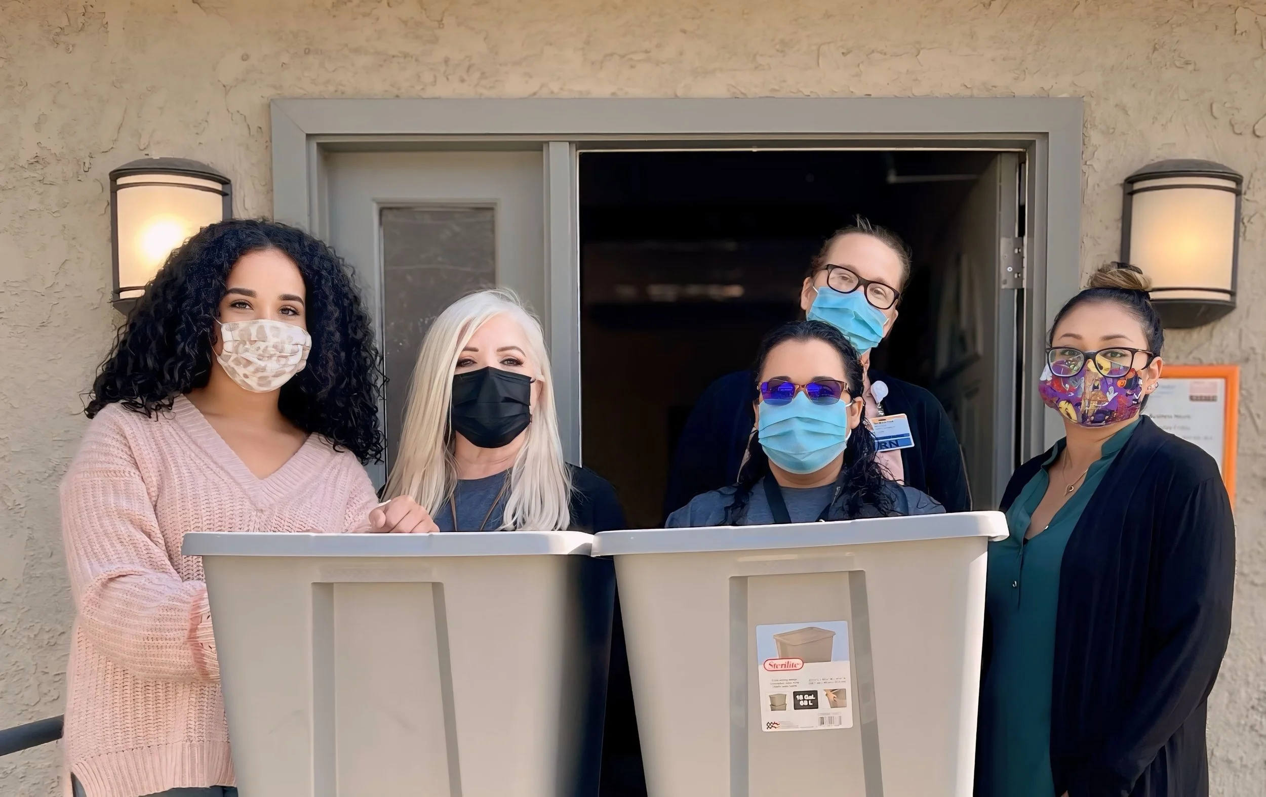 Group of five healthcare workers wearing masks standing outside a building, with one woman holding a large plastic bin