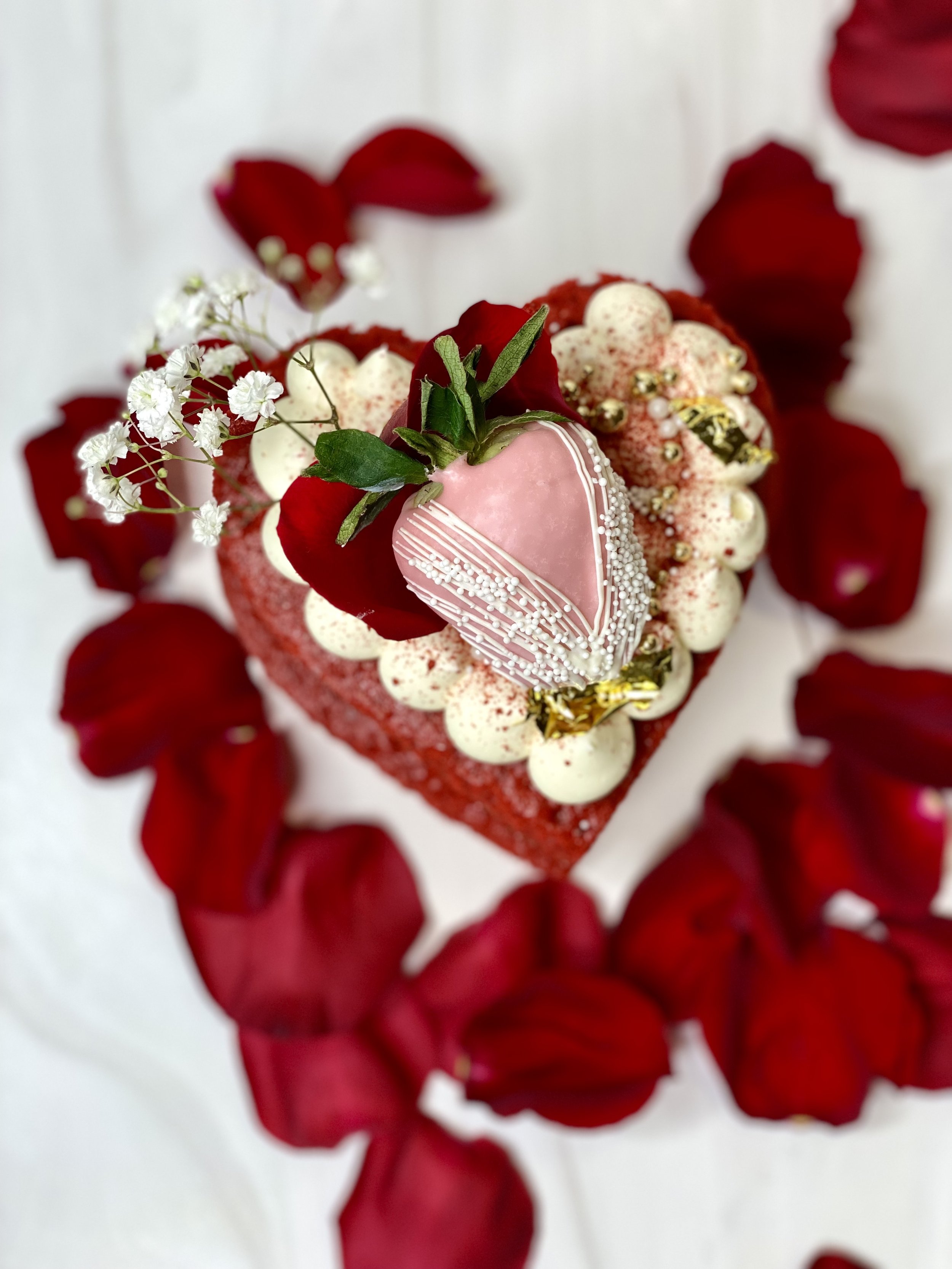 A heart-shaped cake decorated with white and pink frosting, a pink chocolate-dipped strawberry, red rose petals, small white flowers, and gold accents, surrounded by red rose petals.