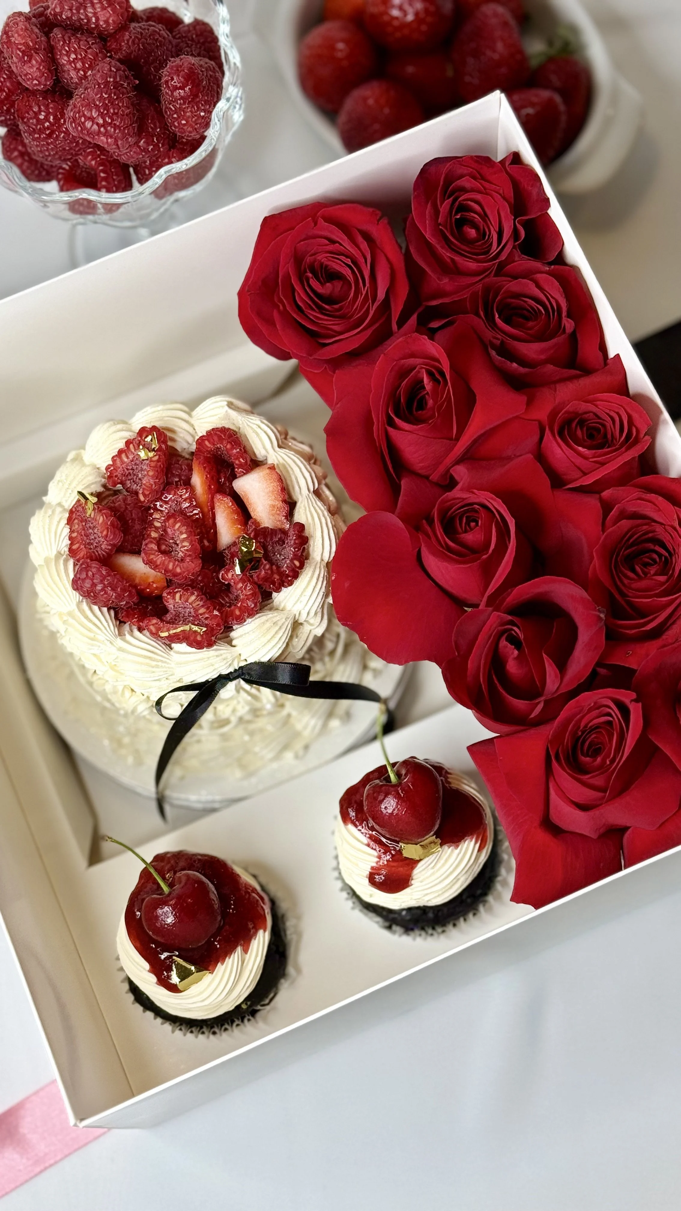 A flower box containing red roses, two cupcakes topped with cherries and whipped cream, and a cake decorated with strawberries and raspberries.
