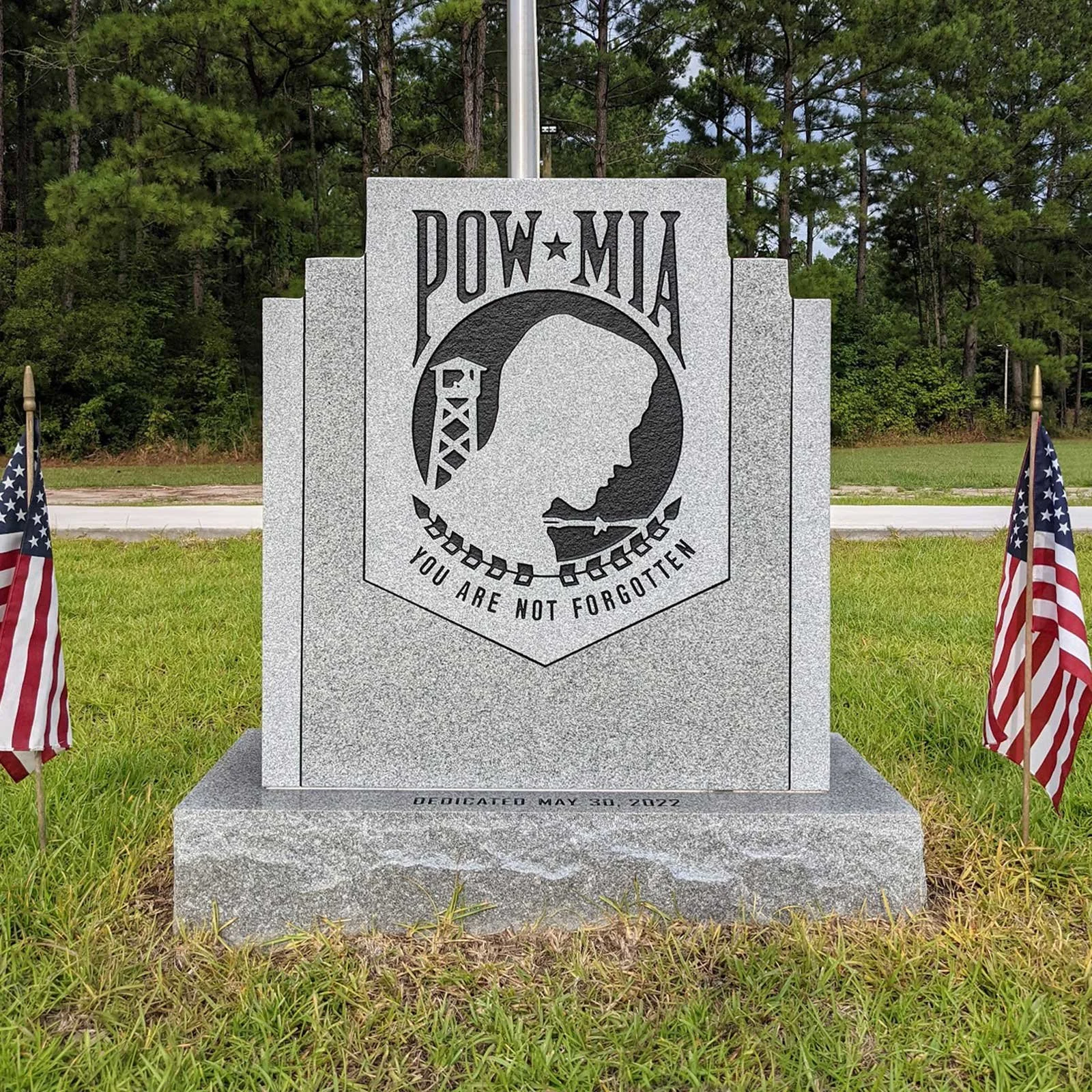 Memorial stone dedicated to POW-MIA with the words 'You Are Not Forgotten' and a silhouette of a soldier's profile, flanked by American flags, in a grassy area with trees in the background.