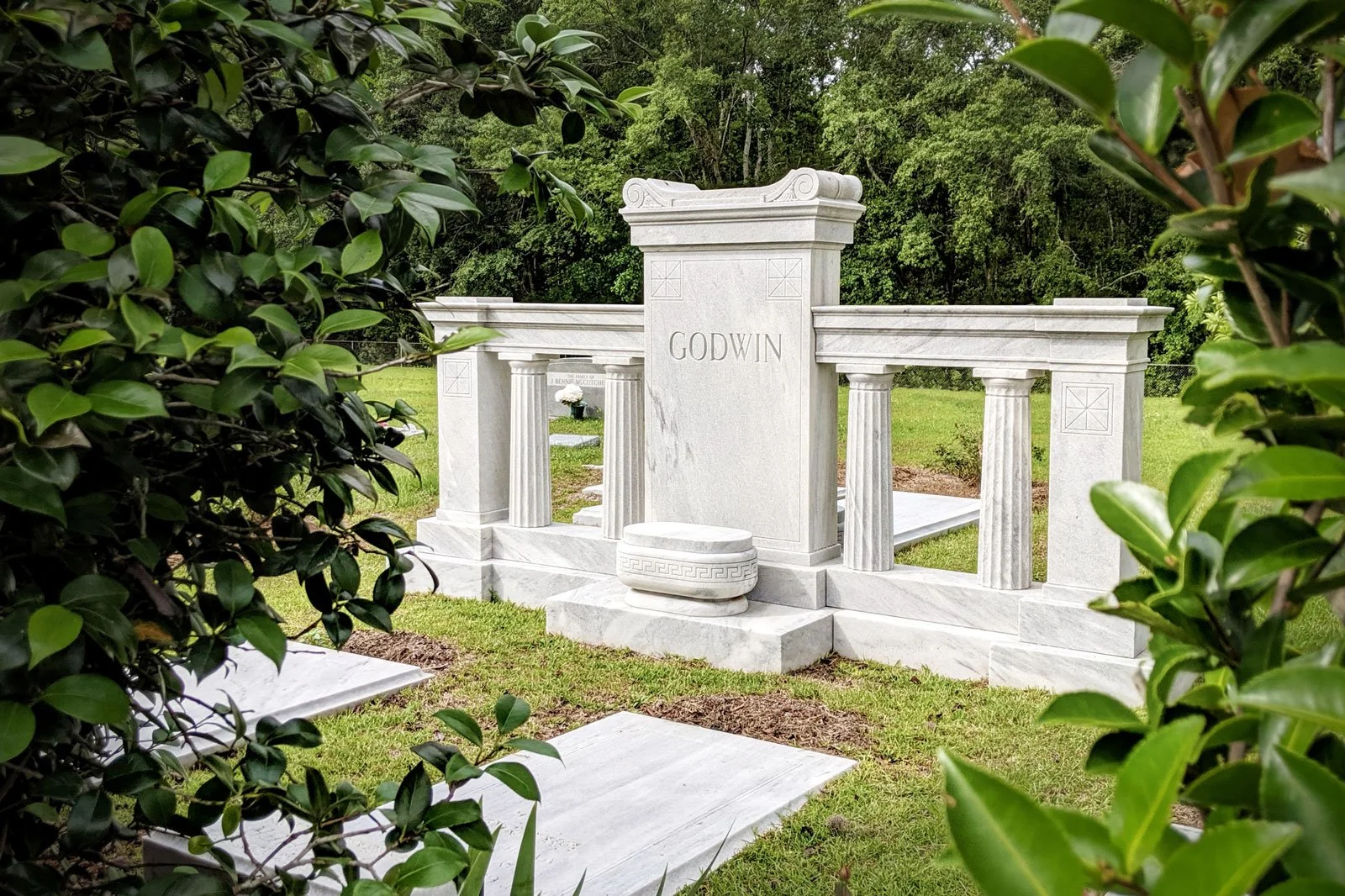 White marble cemetery headstone with the name 'Godwin' engraved, flanked by columns, in a grassy area with trees in the background and green bushes in the foreground.