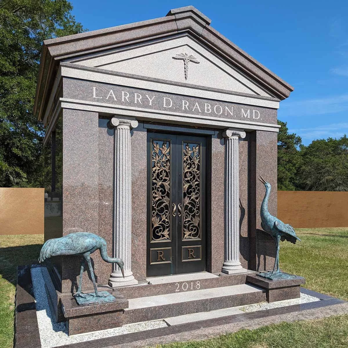 Gravestone with the name Larry D. Rabon, MD, decorated with two crane statues, set in a yard with a green lawn and trees, and a clear blue sky.