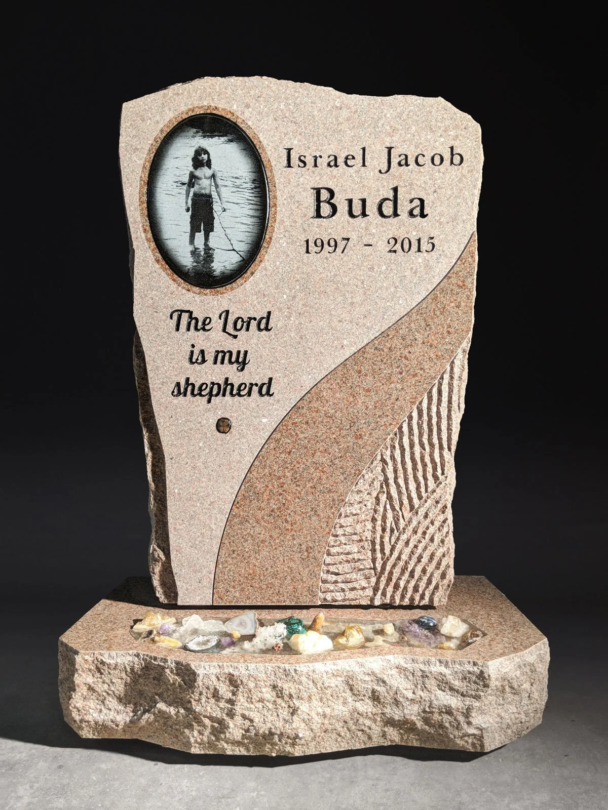 A memorial stone with an engraved photograph of a young boy standing in water, engraved text with the name Israel Jacob Buda, and the message "The Lord is my shepherd," along with the years 1997 to 2015. The stone is decorated with small stones and s