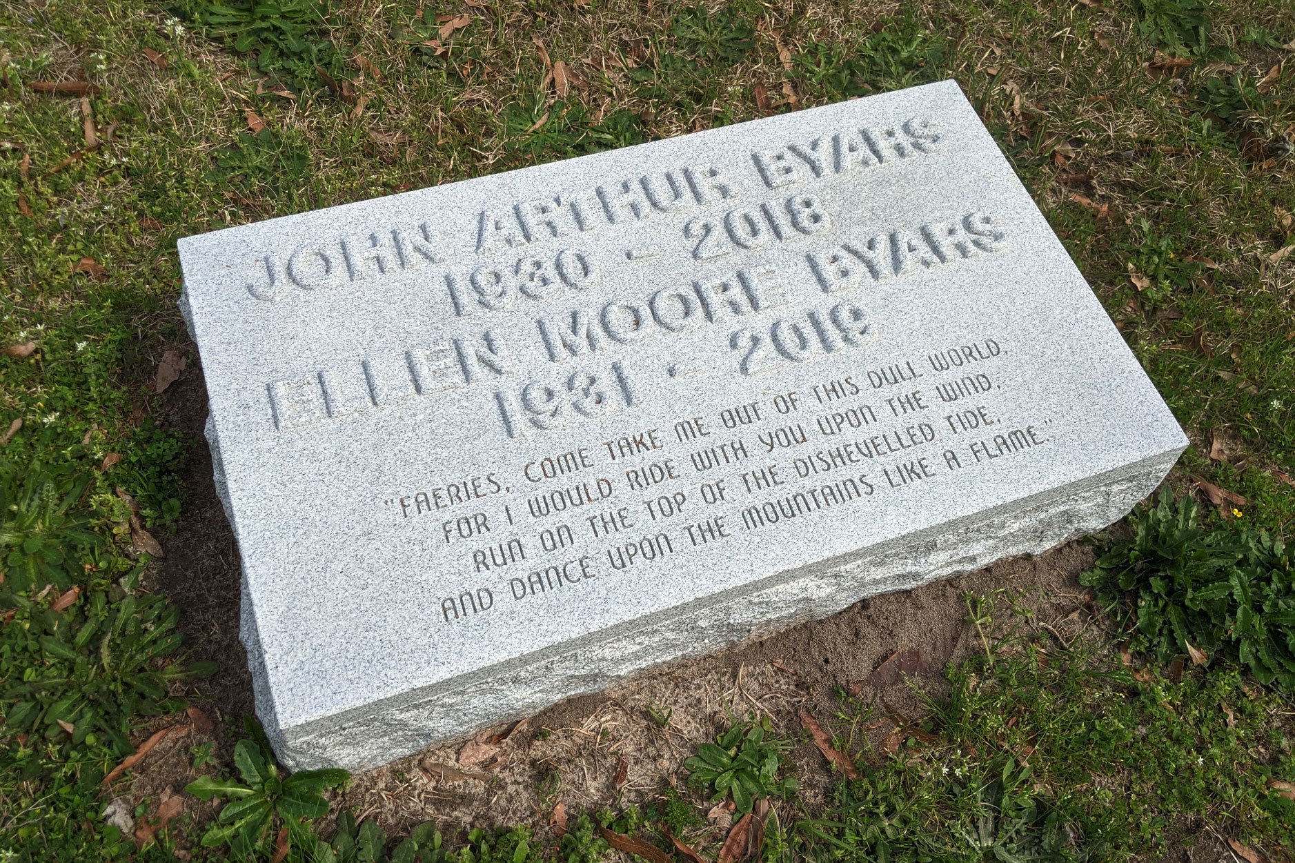 A gray stone memorial gravestone with engraved words and a quote, placed on grass with small green plants around it.